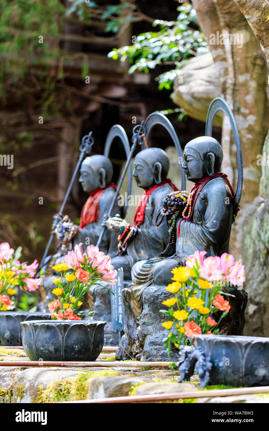 Japan, Miyajima. Daisho-in temple. Line of three Jizo Bosatsu stone statues, two holding scepter, shakujo, and all with red bibs and flowers in front. Stock Photo