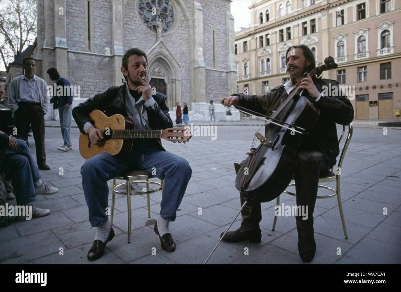 11th May 1993 During the Siege of Sarajevo: the 'cellist of Sarajevo', Vedran Smailović, performs with a guitar-playing friend in front of the Sacred Heart Cathedral. Stock Photo