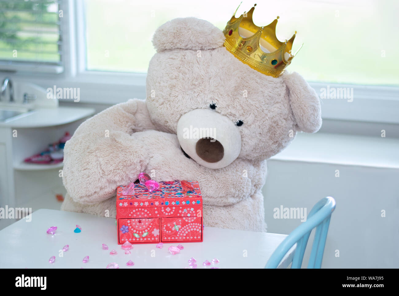 A big teddy bear with a royal crown examines a pile of glittering jewels from his treasure box Stock Photo