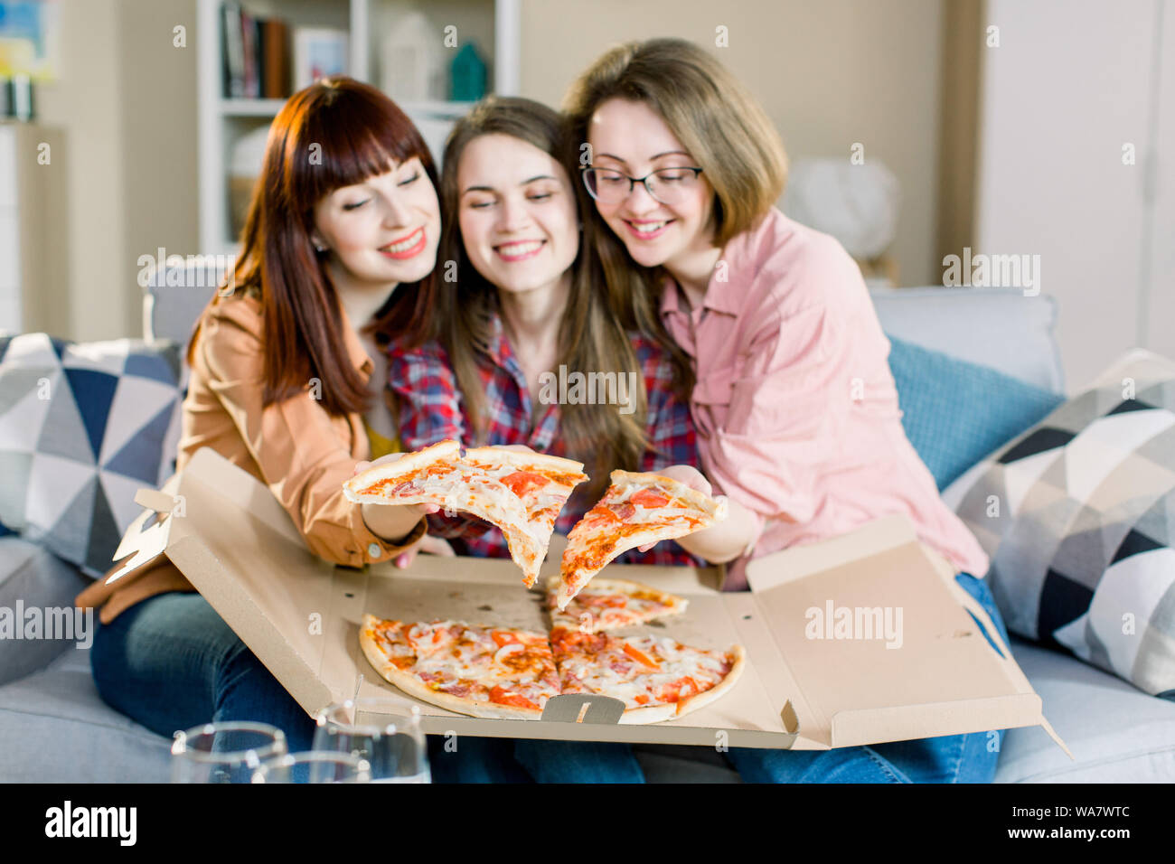 Time to relax! Three stylish girls dressed in blue jeans and shirts eating  pizza with wine on sofa at home Stock Photo - Alamy