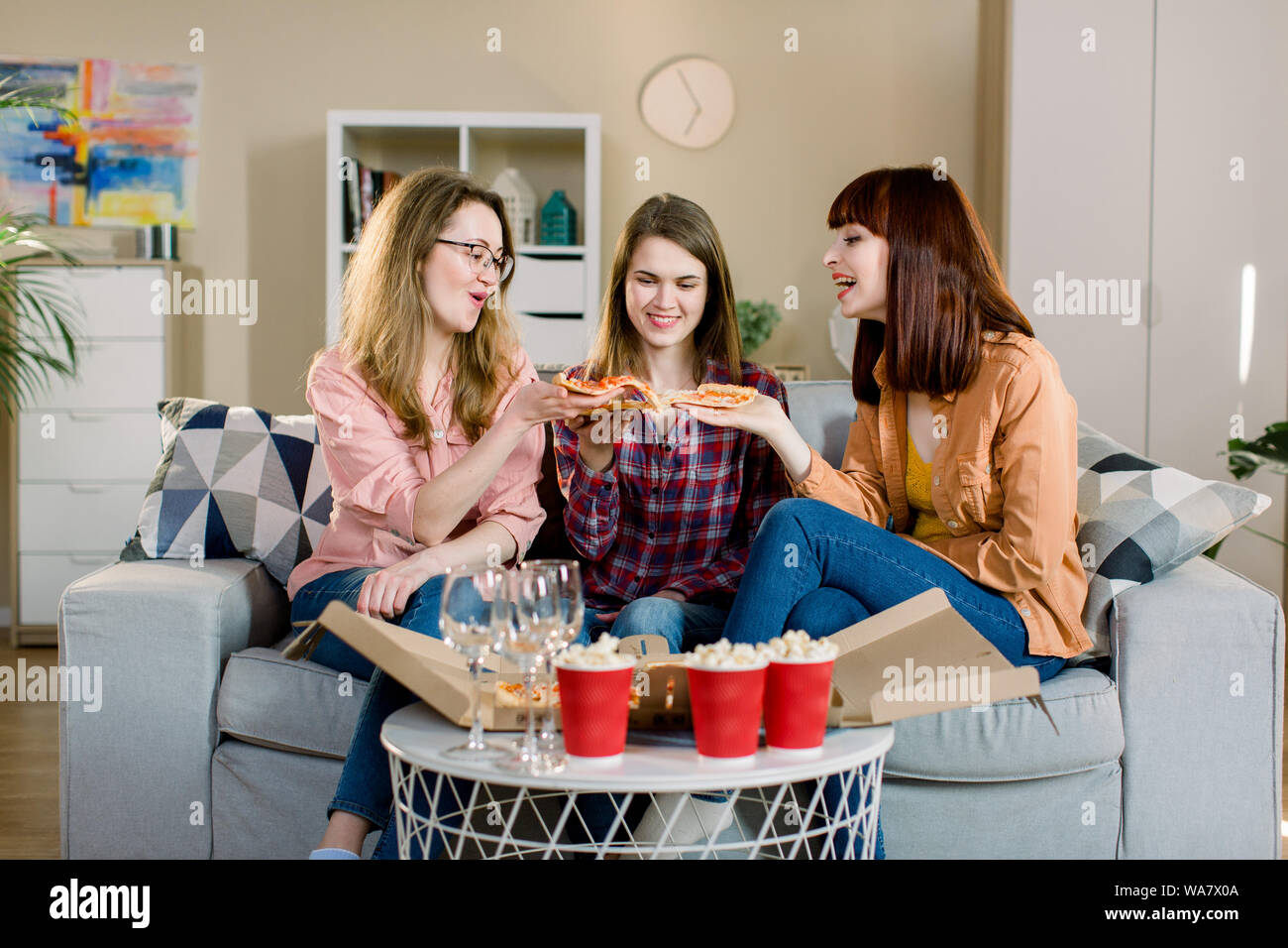 friendship, people, pajama party and junk food concept - happy young three women or girls eating pizza, popcorn and drinking wine at home Stock Photo
