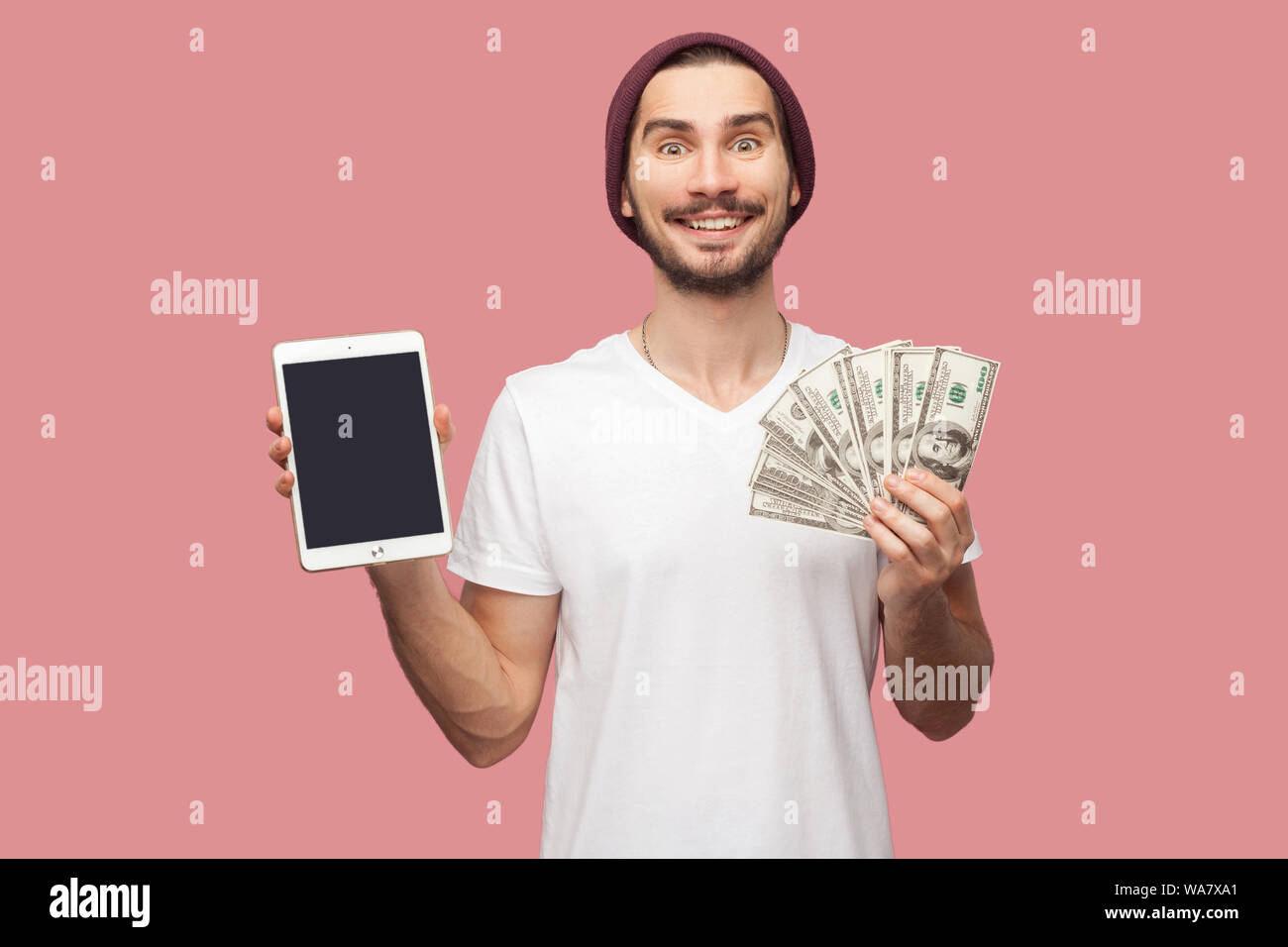 Portrait of happy handsome bearded young hipster man in white shirt and casual hat standing, holding empty screen ipad and green present box. Indoor, Stock Photo