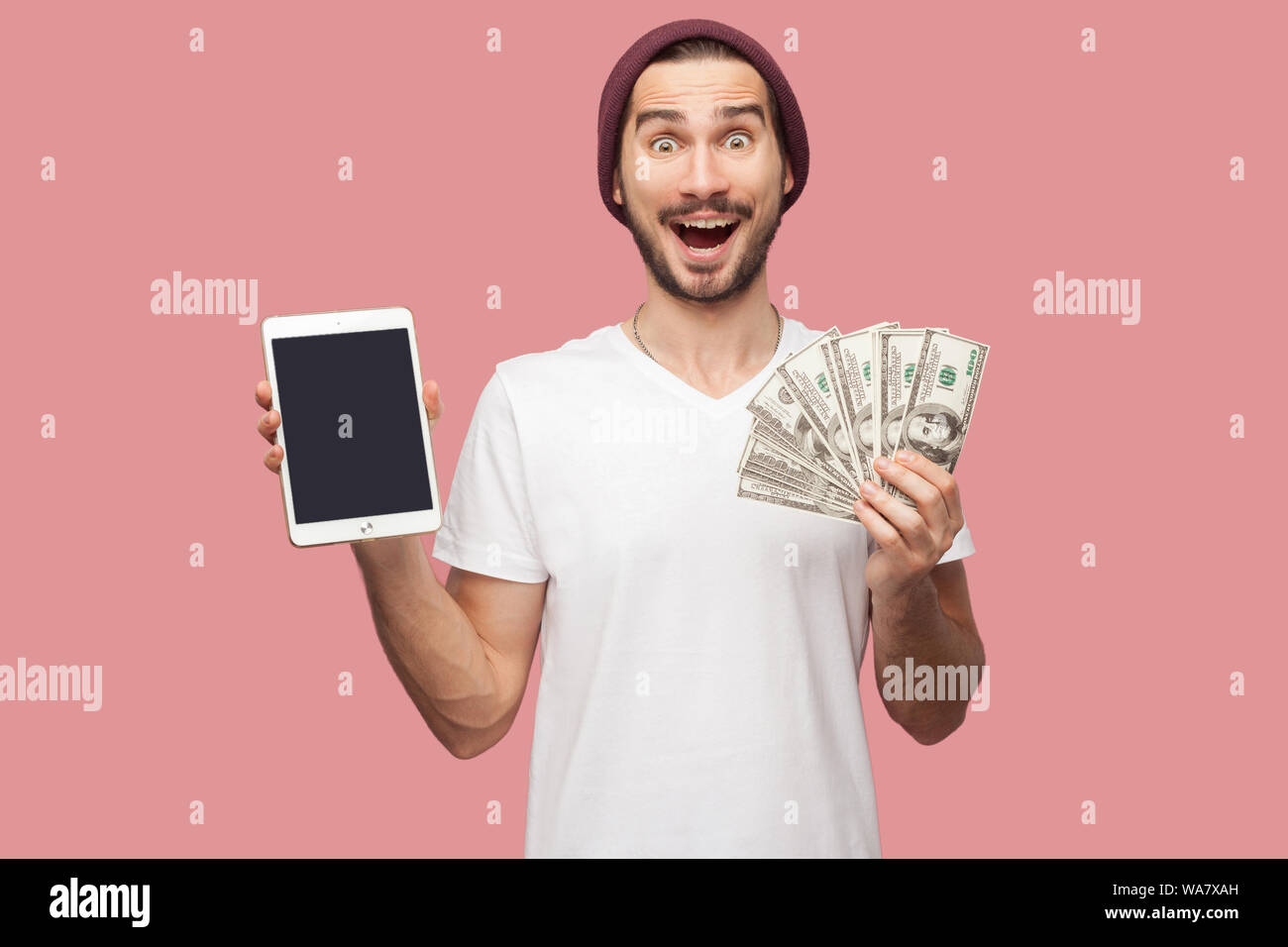 Portrait of suprised handsome bearded young hipster man in white shirt and casual hat standing, holding empty screen ipad and green present box. Indoo Stock Photo