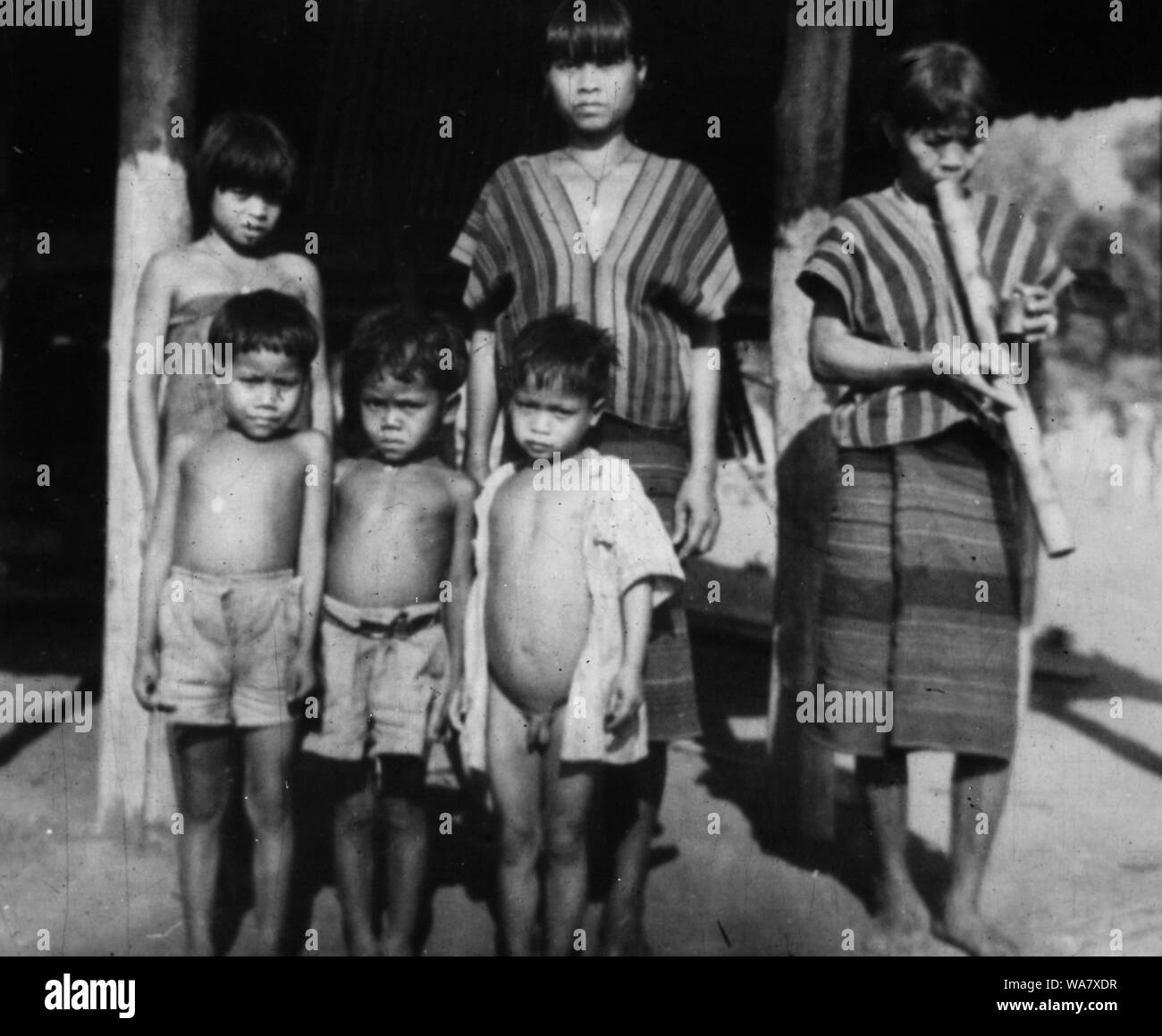 AJAXNETPHOTO. 1953-1957 (APPROX). INDO CHINA. VIETNAM. (IN-COUNTRY LOCATION UNKNOWN.) -  GROUP OF GIRLS, ONE SMOKING A BAMBOO WATER PIPE AND YOUNG BOYS POSE FOR THE CAMERA OUTSIDE HOUSE ON STILTS. PHOTO:JEAN CORRE/AJAXREF:RX7 191508 242 Stock Photo