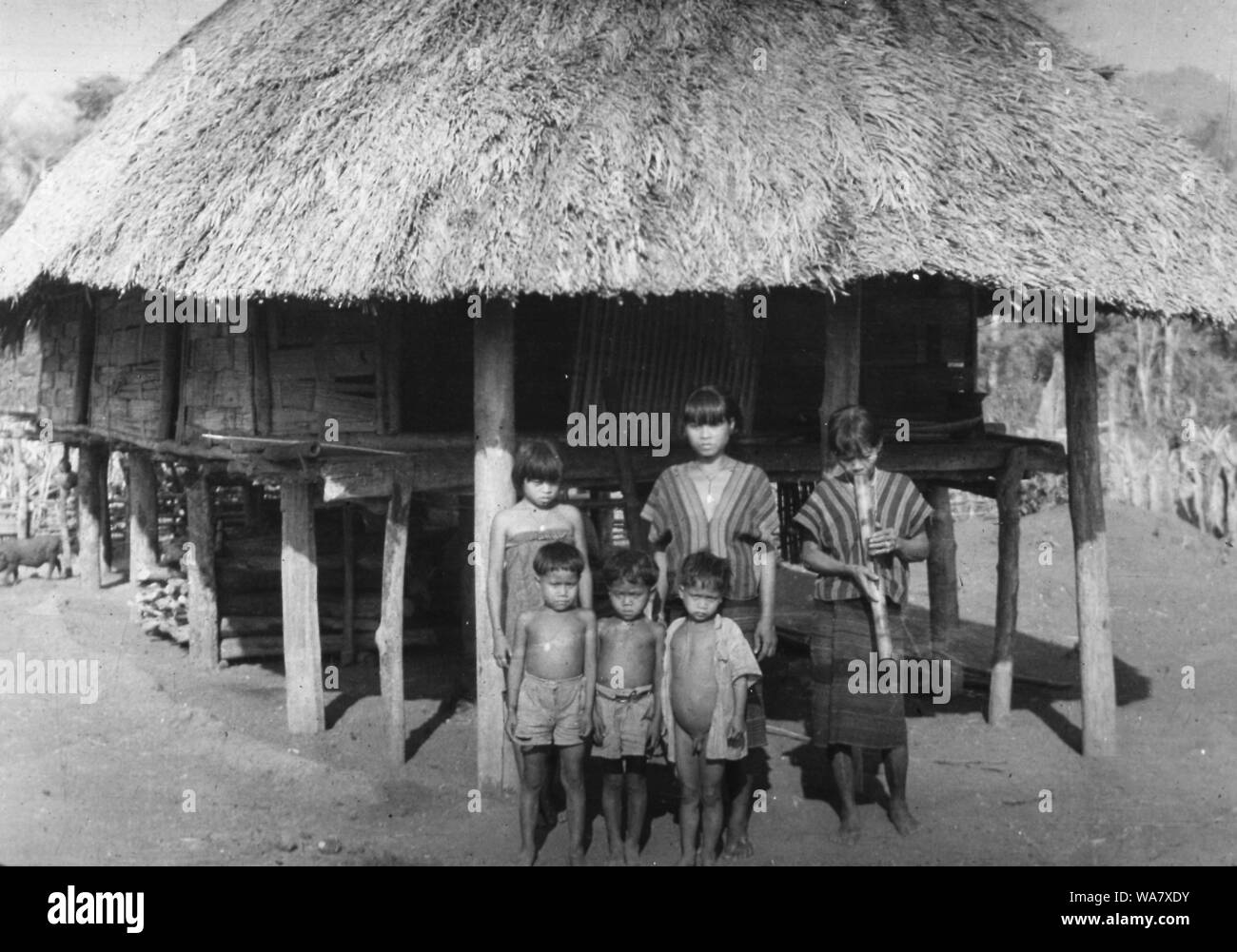 AJAXNETPHOTO. 1953-1957 (APPROX). INDO CHINA. VIETNAM. (IN-COUNTRY LOCATION UNKNOWN.) -  GIRLS, ONE SMOKING A TRADITIONAL BAMBOO WATER PIPE, AND YOUNG BOYS POSE FOR THE CAMERA OUTSIDE THATCHED ROOF HOUSE (HOOCH) ON STILTS. PHOTO:JEAN CORRE/AJAXREF:RX7 191508 243 Stock Photo