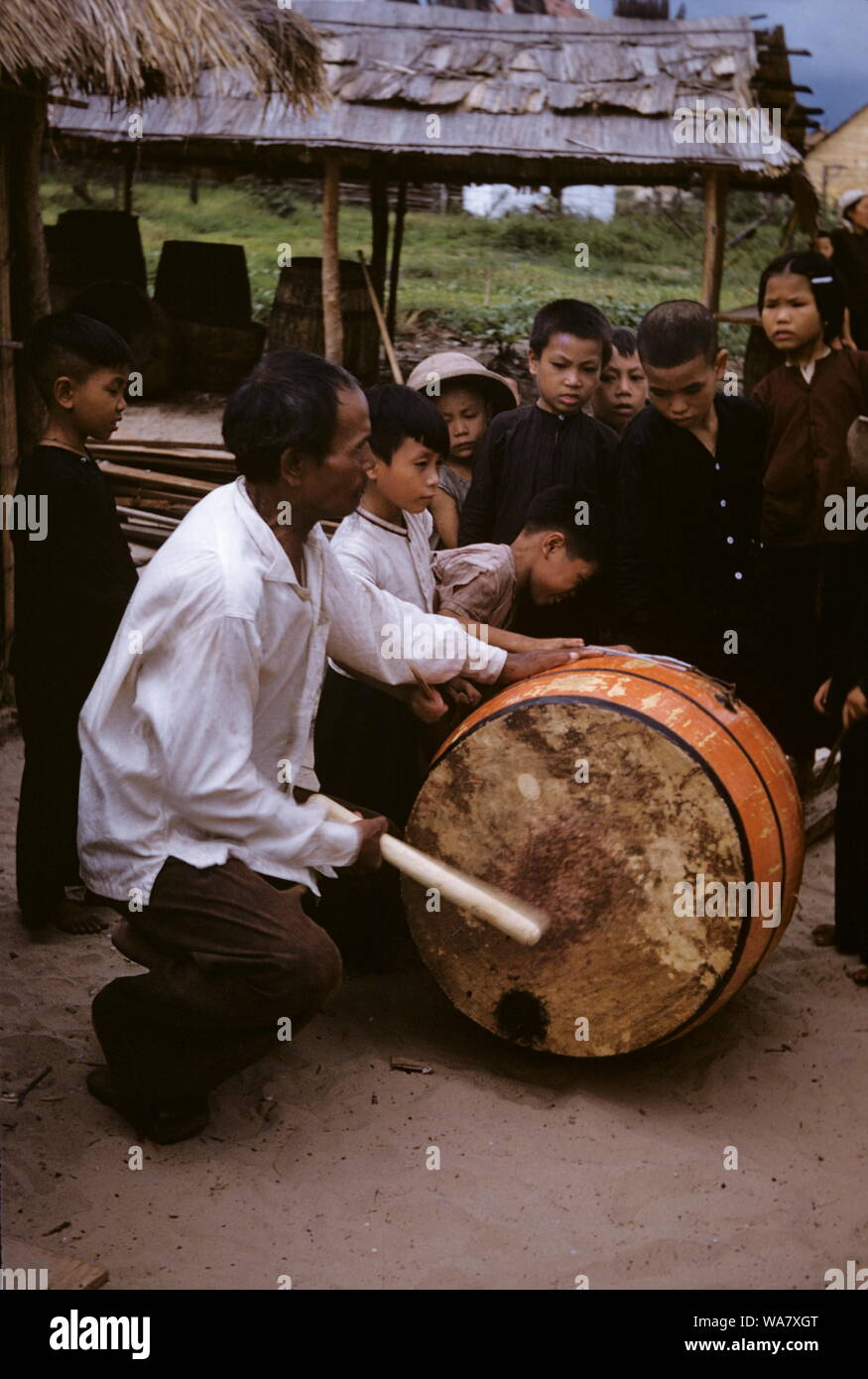 AJAXNETPHOTO. 1955 (APPROX). INDO CHINA. VIETNAM. (IN-COUNTRY LOCATION UNKNOWN.) - BEATING THE DRUM - YOUNG PEASANT CHILDREN LOOK ON AS A MAN BANGS A DRUM. PHOTO:JEAN CORRE/AJAXREF:JC1955 Stock Photo