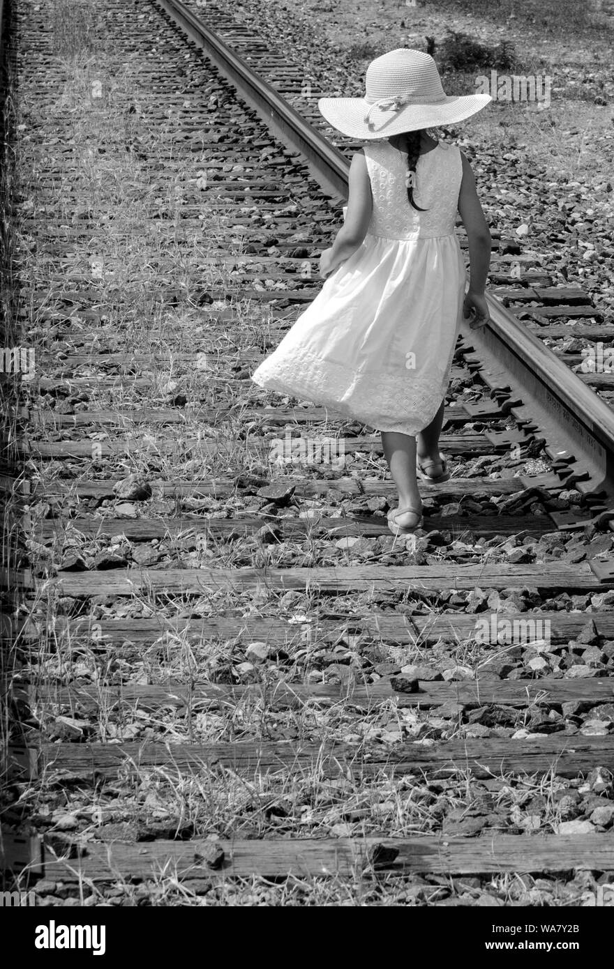 black and white of a little girl in a floppy hat and white sundress walking along a rail road track Stock Photo