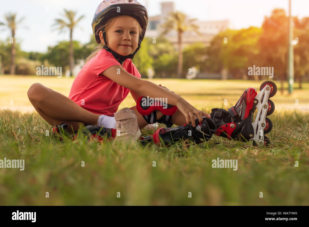 Cute little blonde girl sitting on green grass and putting on roller skates Stock Photo