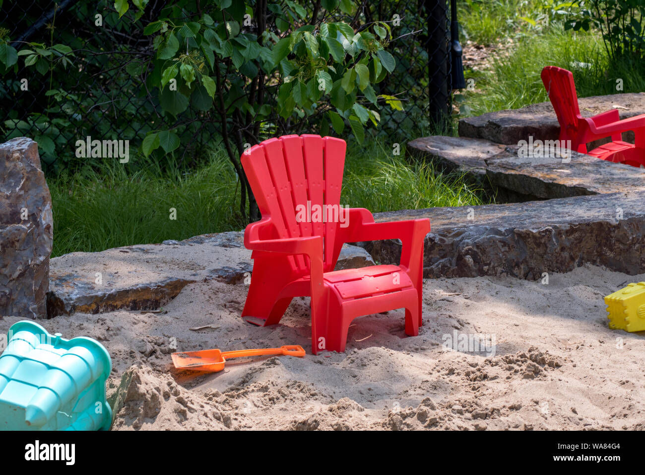 small child's Adirondack chair rests in a big outdoor sand box with plenty of sand toys Stock Photo