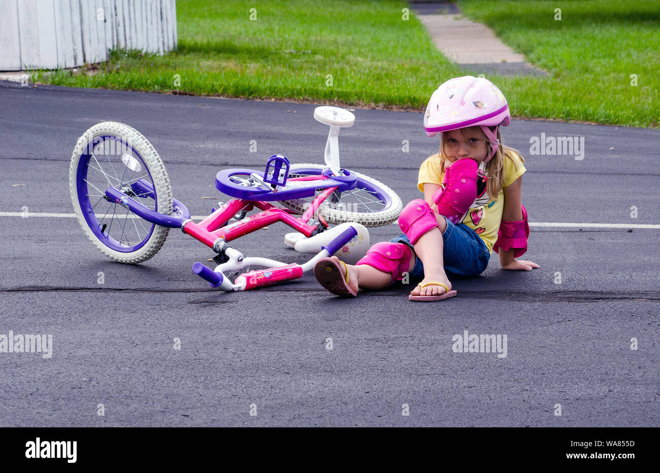 Little girl sits on the ground in helmet and safety gear, after falling off her bike, while learning to ride with training wheels Stock Photo