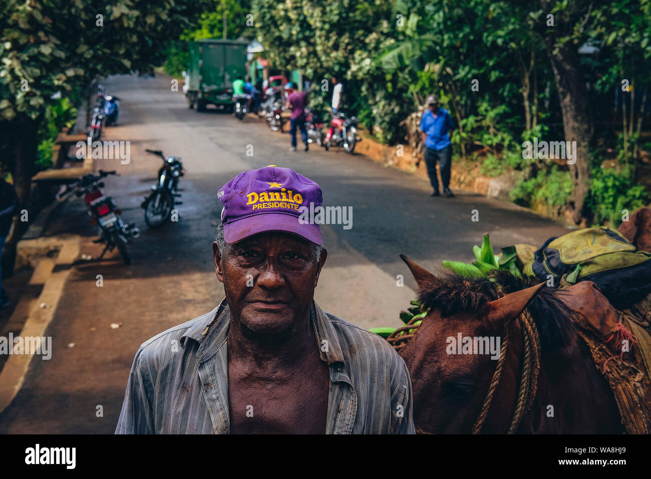 Old man from the countryside is seen in a village street wearing a cap that states 'Danilo Presidente' near a loaded donkey Stock Photo