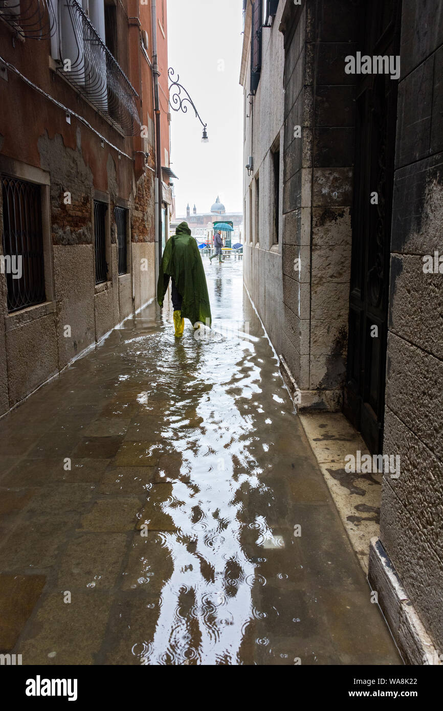A caped tourist, on a rainy day, walking through a flooded narrow street during an Acqua alta (high water) event, Calle de la Rasse, Venice, Italy Stock Photo
