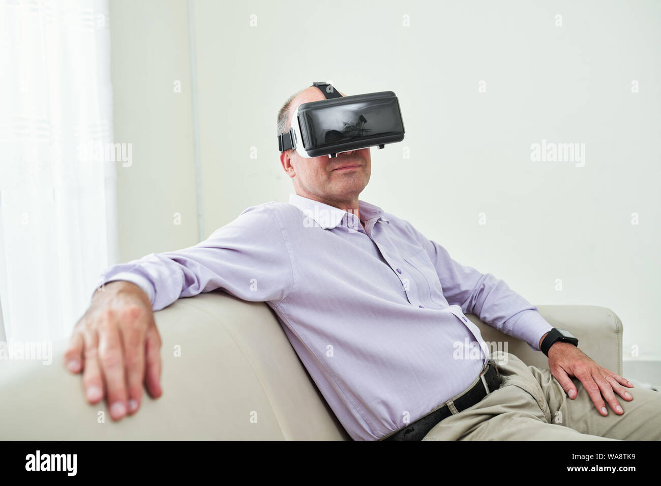 Portrait of smiling mature entrepreneur in lavender shirt resting on sofa in virtual reality glasses Stock Photo