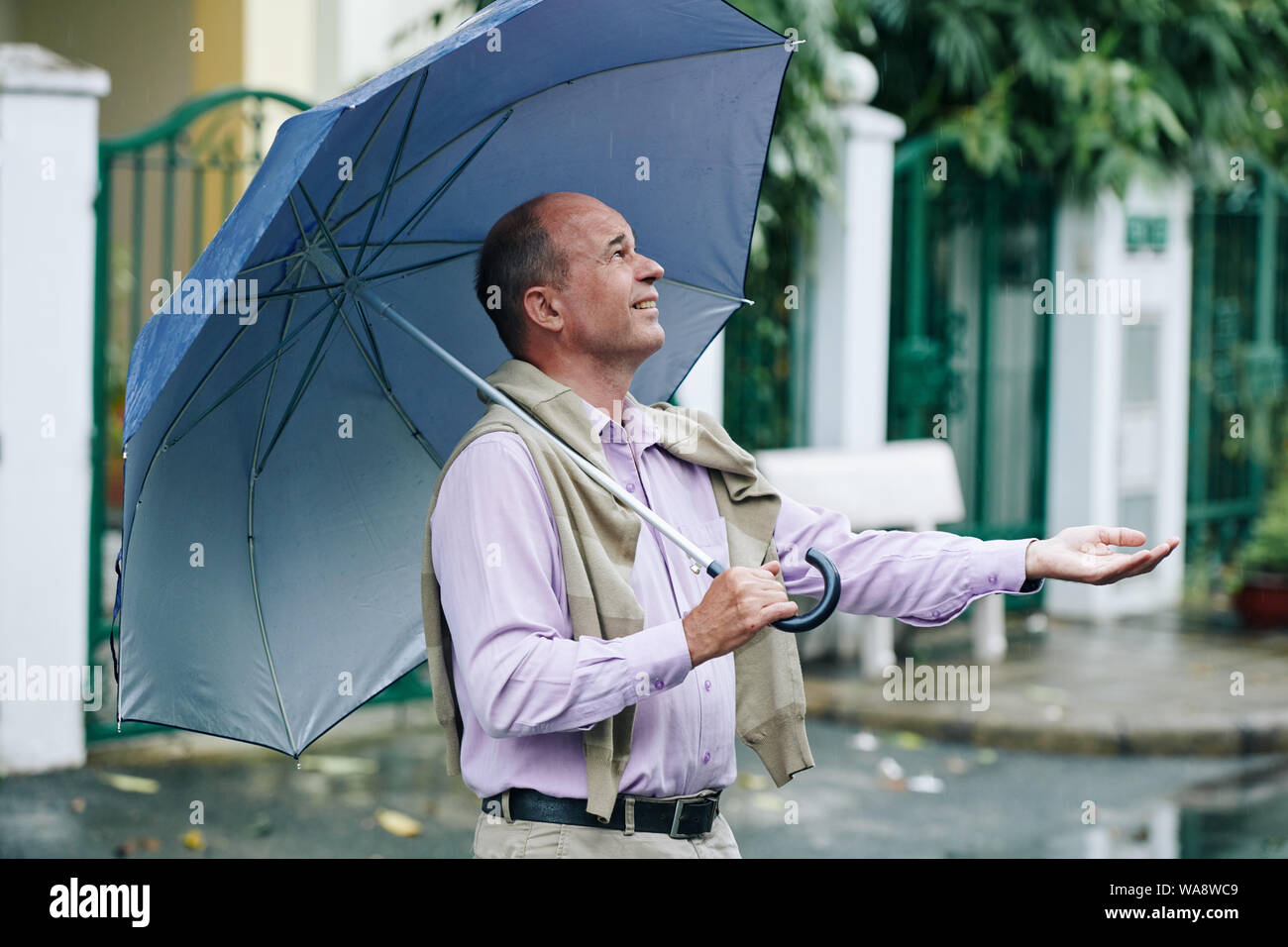 Happy middle-aged man with umbrella outstretching hand to catch rain drops Stock Photo