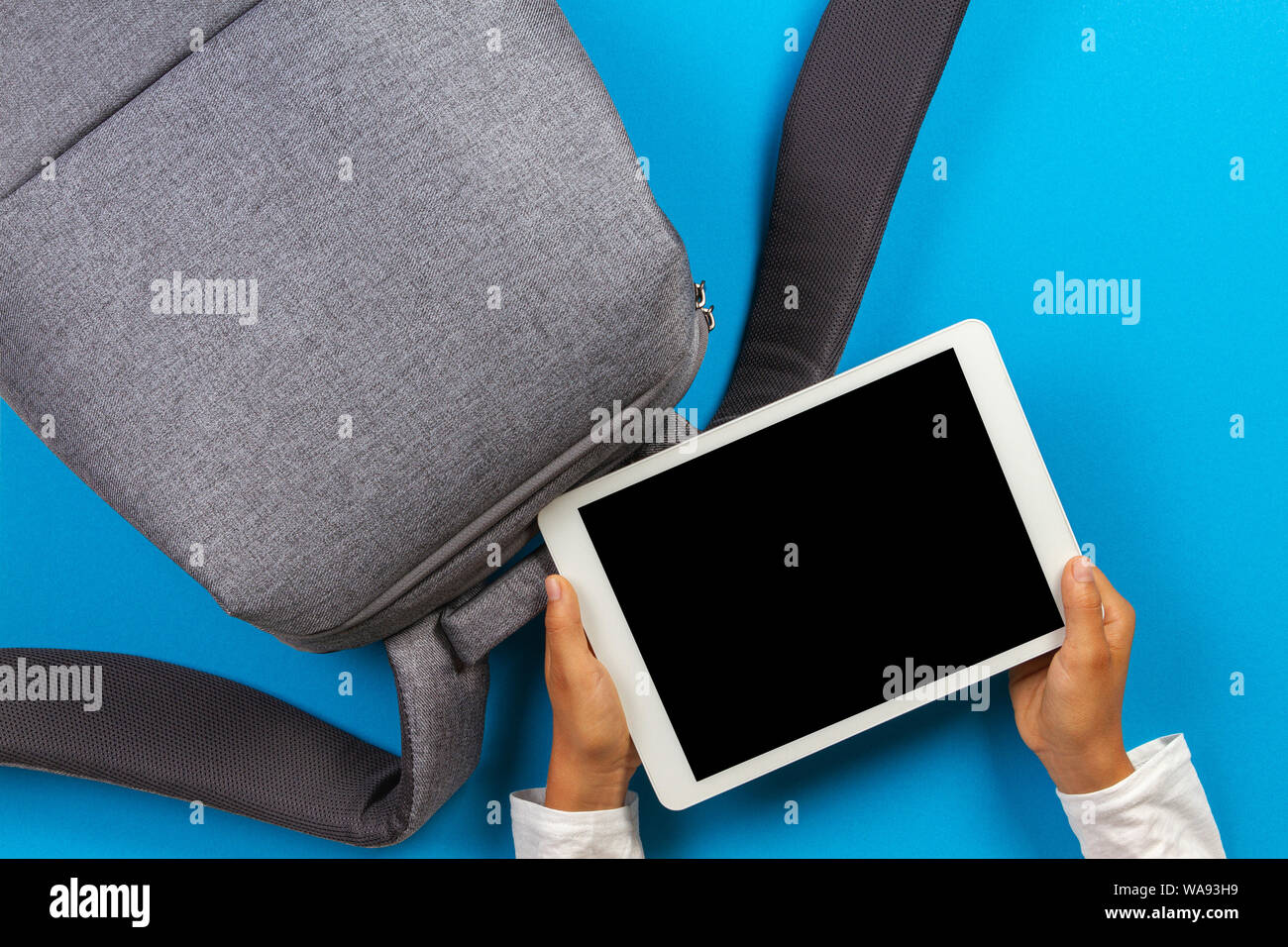 Kid hands with tablet computer and gray backpack over light blue background Stock Photo
