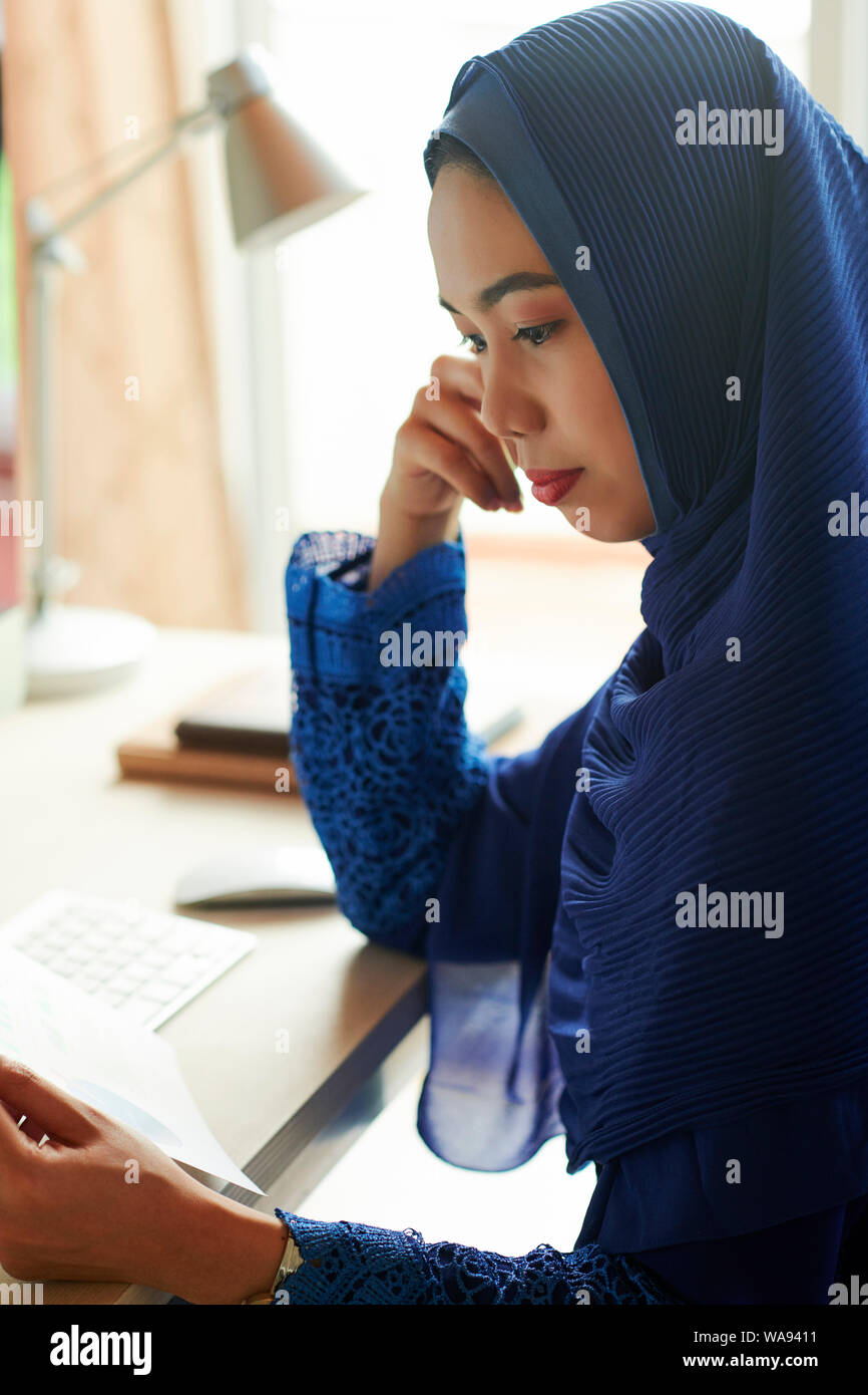 Pensive young muslim woman in hijab sitting at desk and reading ...