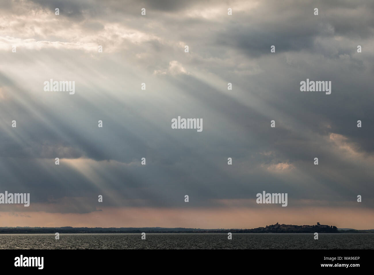 Sunrays at near sunset, with dark clouds in the background, an orange sky, and Trasimeno lake (Umbria, Italy) below Stock Photo