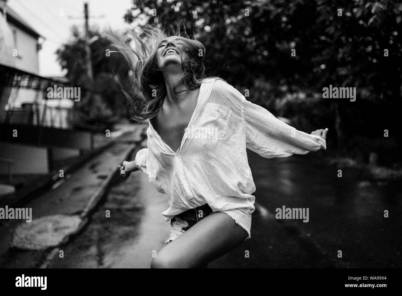 Beautiful lady enjoys fresh rai, she walking outdoor in wet clothes and laughing. White big shirt on her sexy body, short jeans. Tanned soft skin, hot Stock Photo