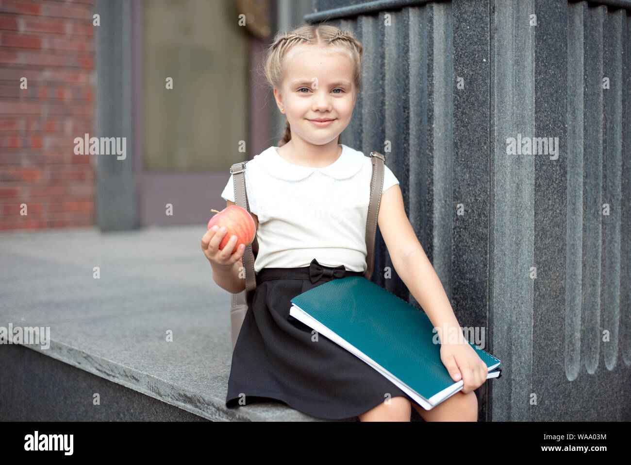 Happy little schoolgirl with blond hair in school uniform with lunch, book sits near school. Stock Photo