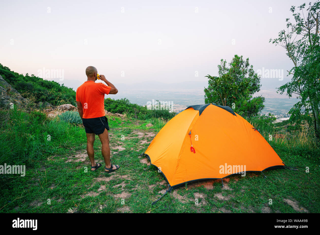 Man is drinking from cup near an orange tent standing on hill Stock Photo