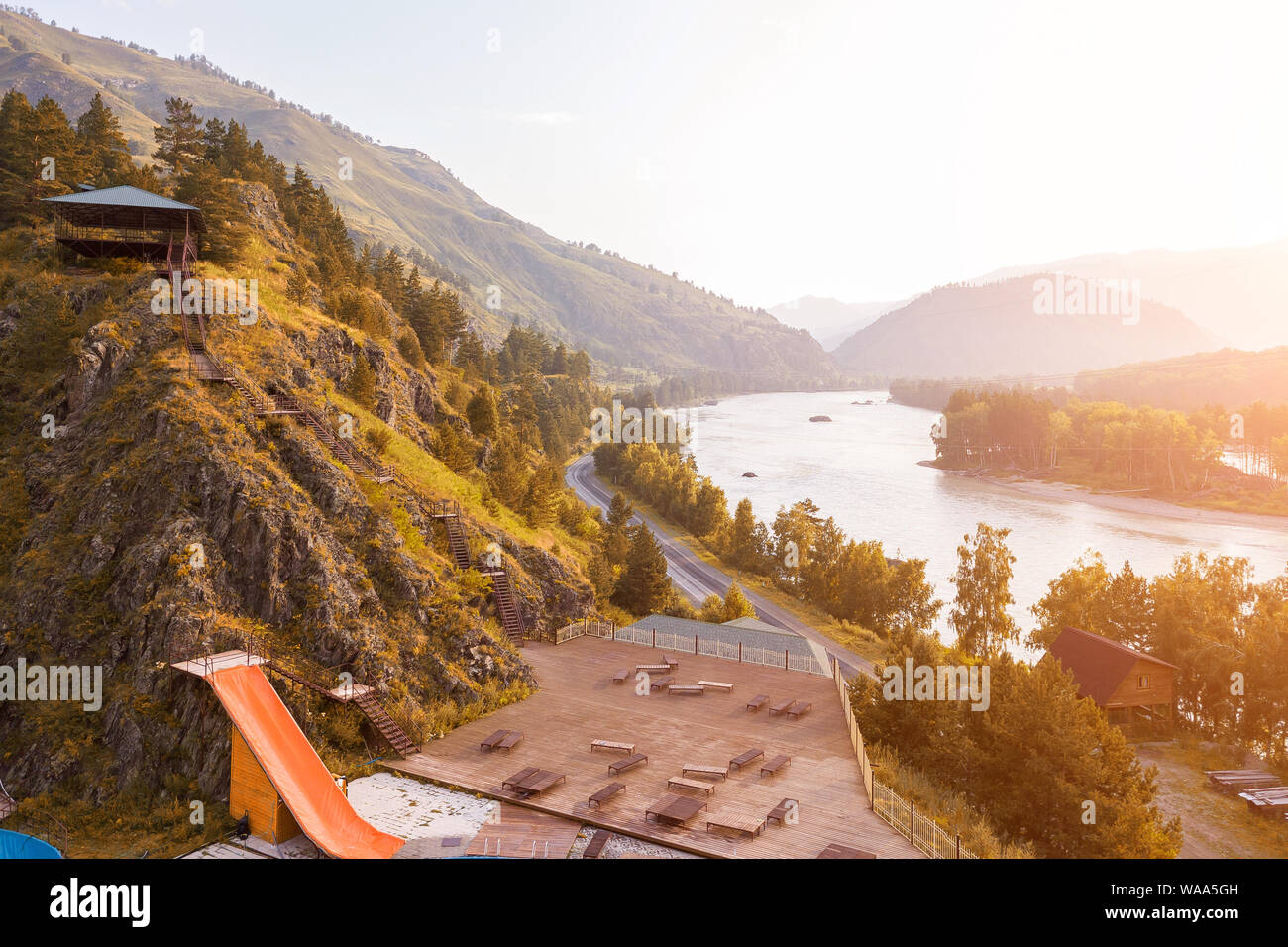 outdoor pool in the mountains with a yellow water slide attraction with wooden chaise-longue, blue water, separated by a metal fence, overlooking the Stock Photo