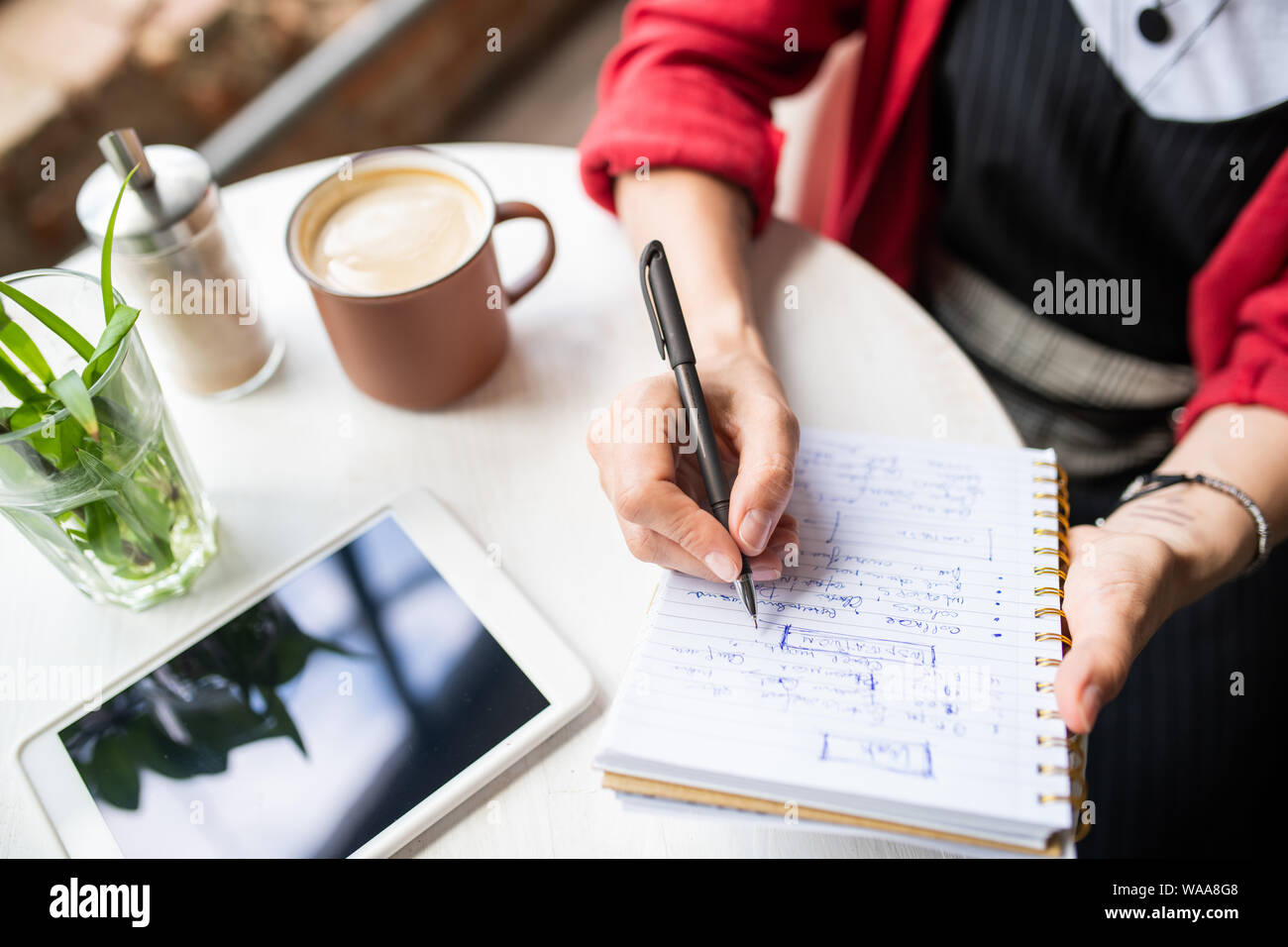 Young woman writing down working notes in notebook while sitting by table Stock Photo