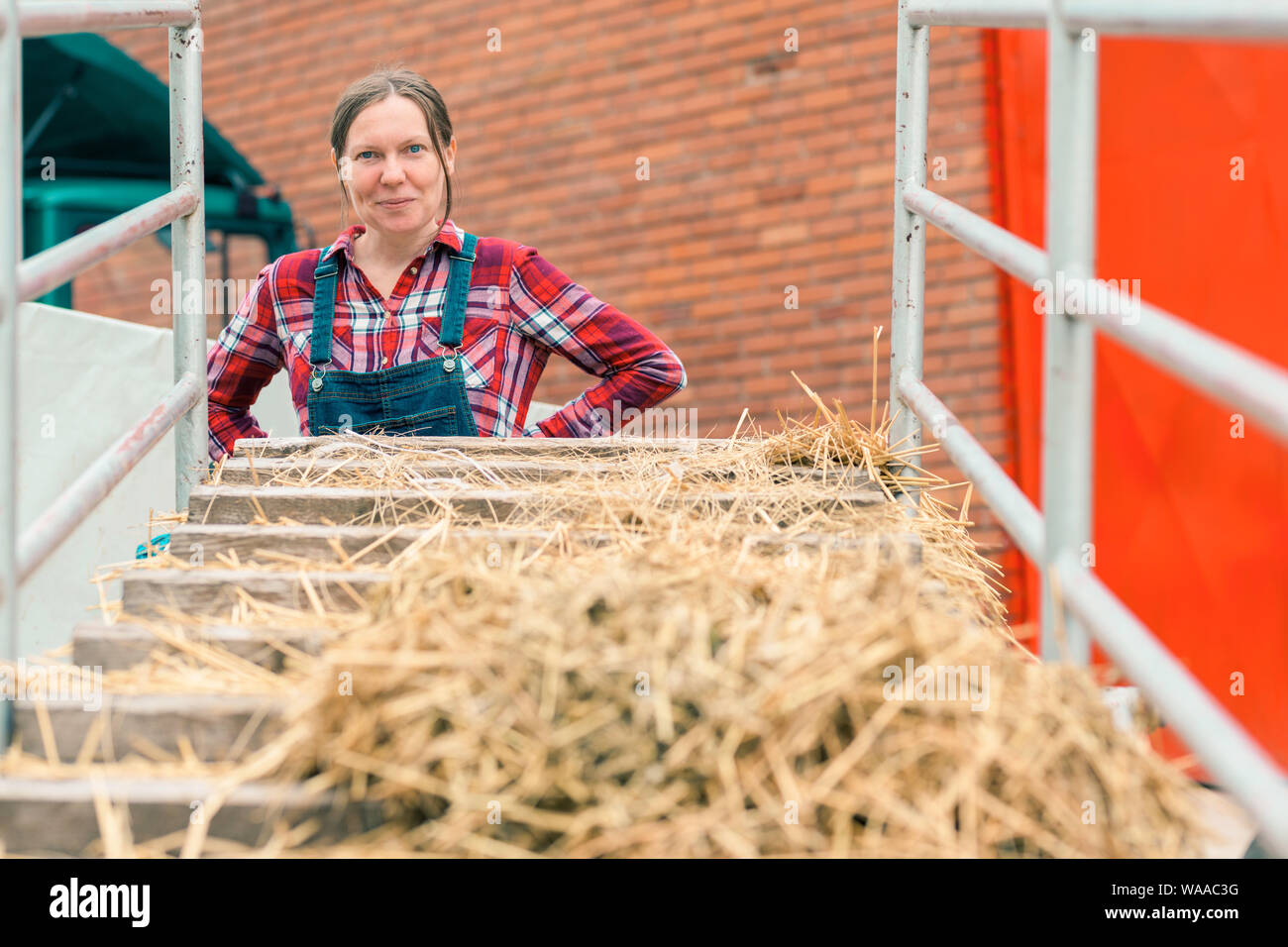 Satisfied happy female farmer with small hay wagon on agricultural farm Stock Photo
