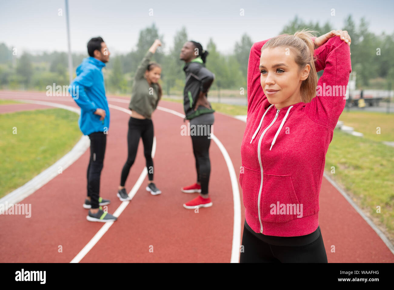 Happy young blonde sportswoman doing warming up exercise on stadium Stock Photo