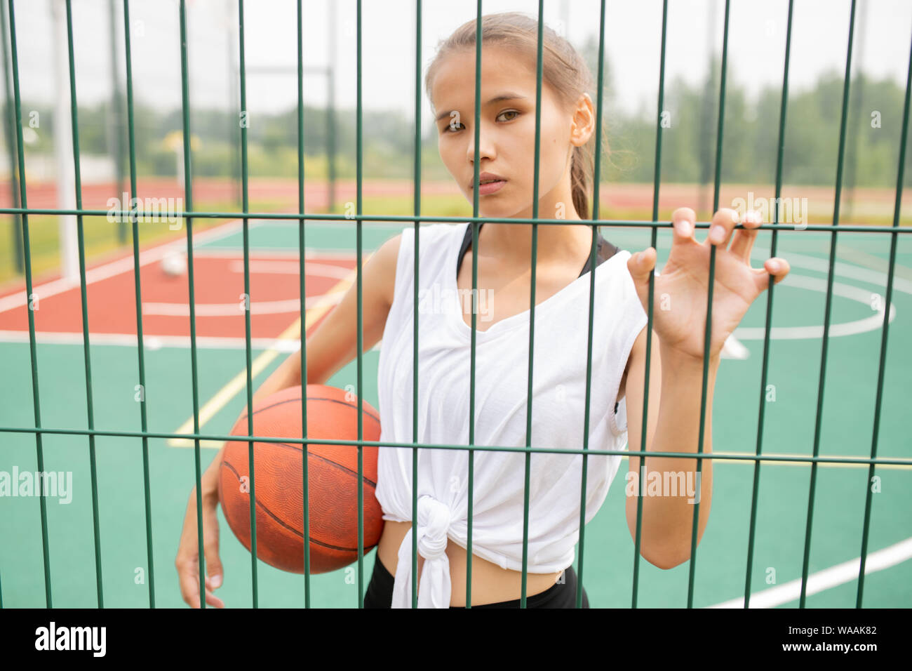 Pretty girl in activewear holding ball while looking at you through sports net Stock Photo