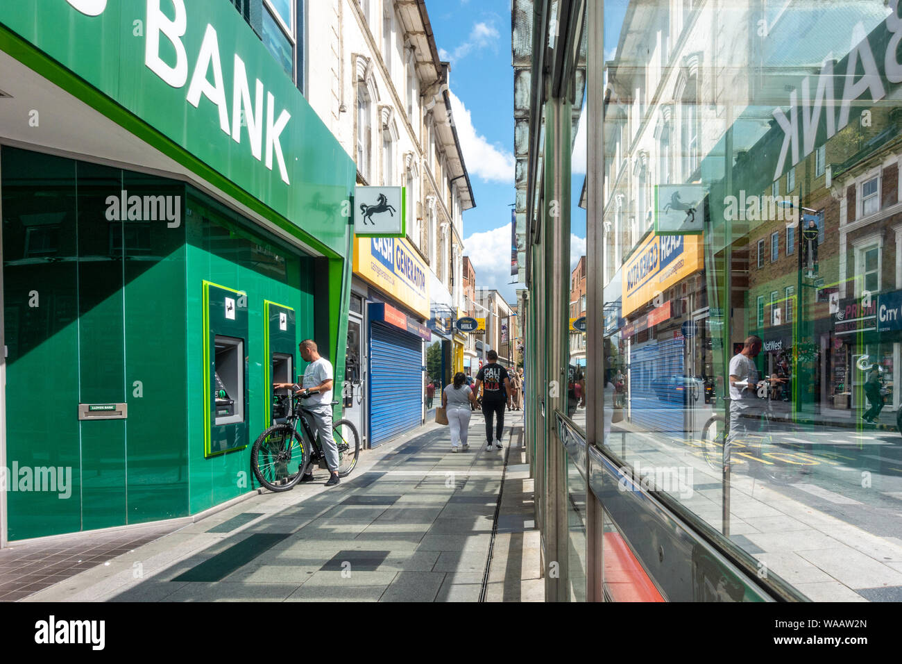 A man uses an ATM machine at the Slough High Street branch of Lloyds bank. The scene is seen reflected in a glass bus stop. Stock Photo