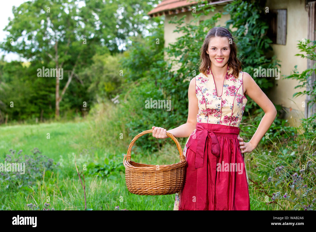 portrait of young woman in dirndl standing at farmhouse and holding basket Stock Photo
