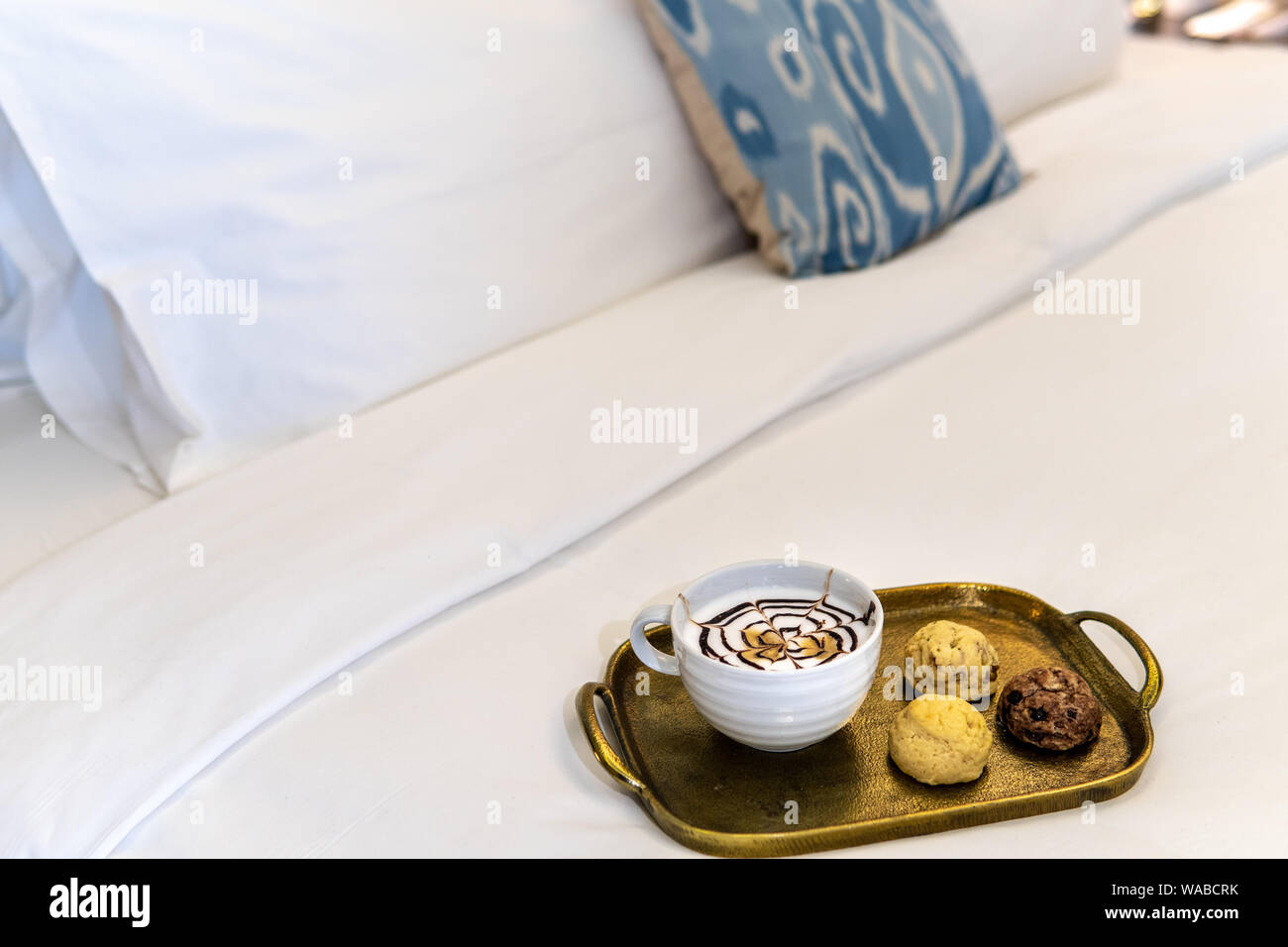 cappuccino with fancy chocolate design on top with trhee various baked cookies on gold tray in hotel room on bed Stock Photo