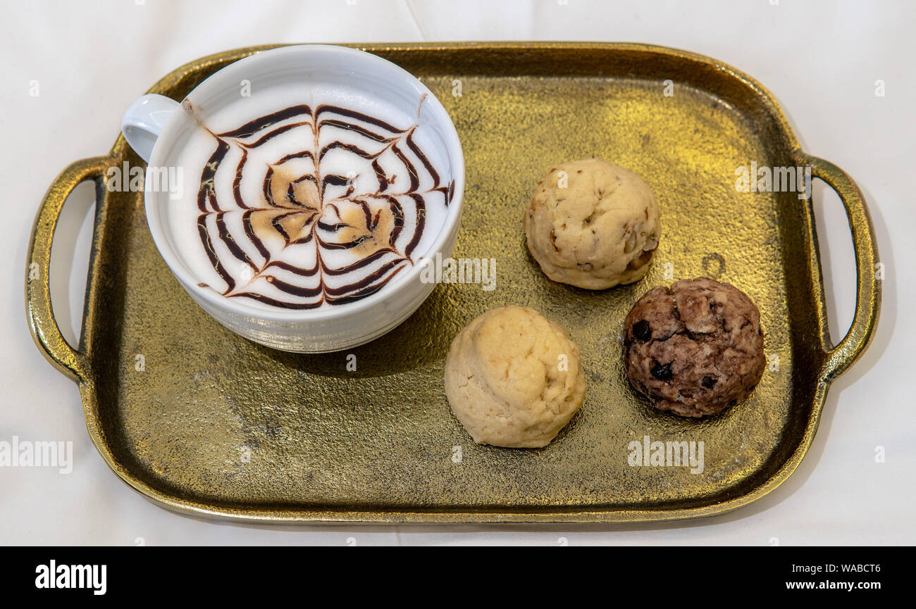 cappuccino with fancy chocolate design on top with trhee various baked cookies on gold tray in hotel room on bed Stock Photo