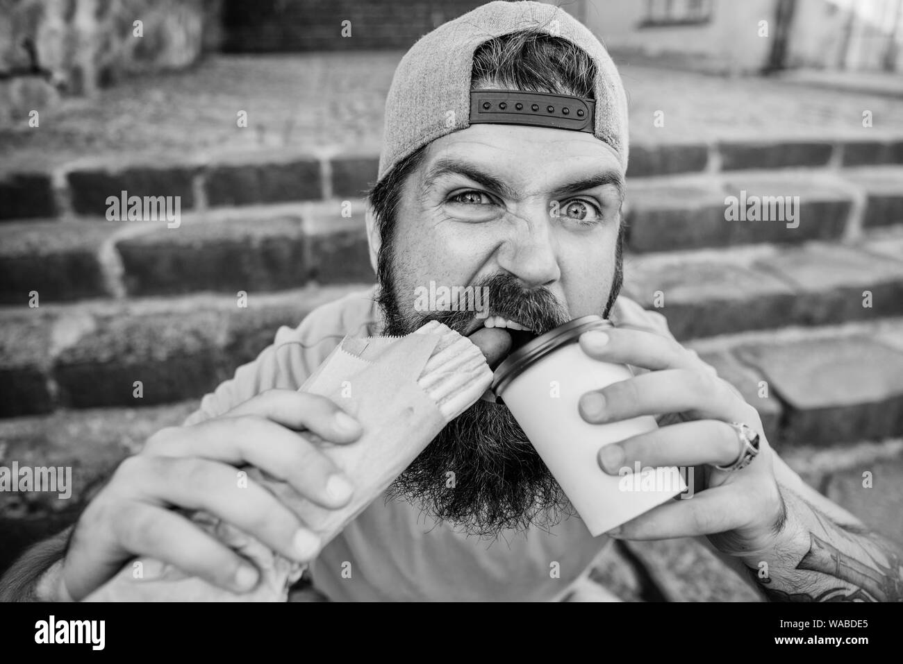 Carefree hipster eat junk food while sit stairs. Snack for good mood. Guy eating hot dog. Street food concept. Man bearded eat tasty sausage and drink paper cup. Urban lifestyle nutrition. Junk food. Stock Photo