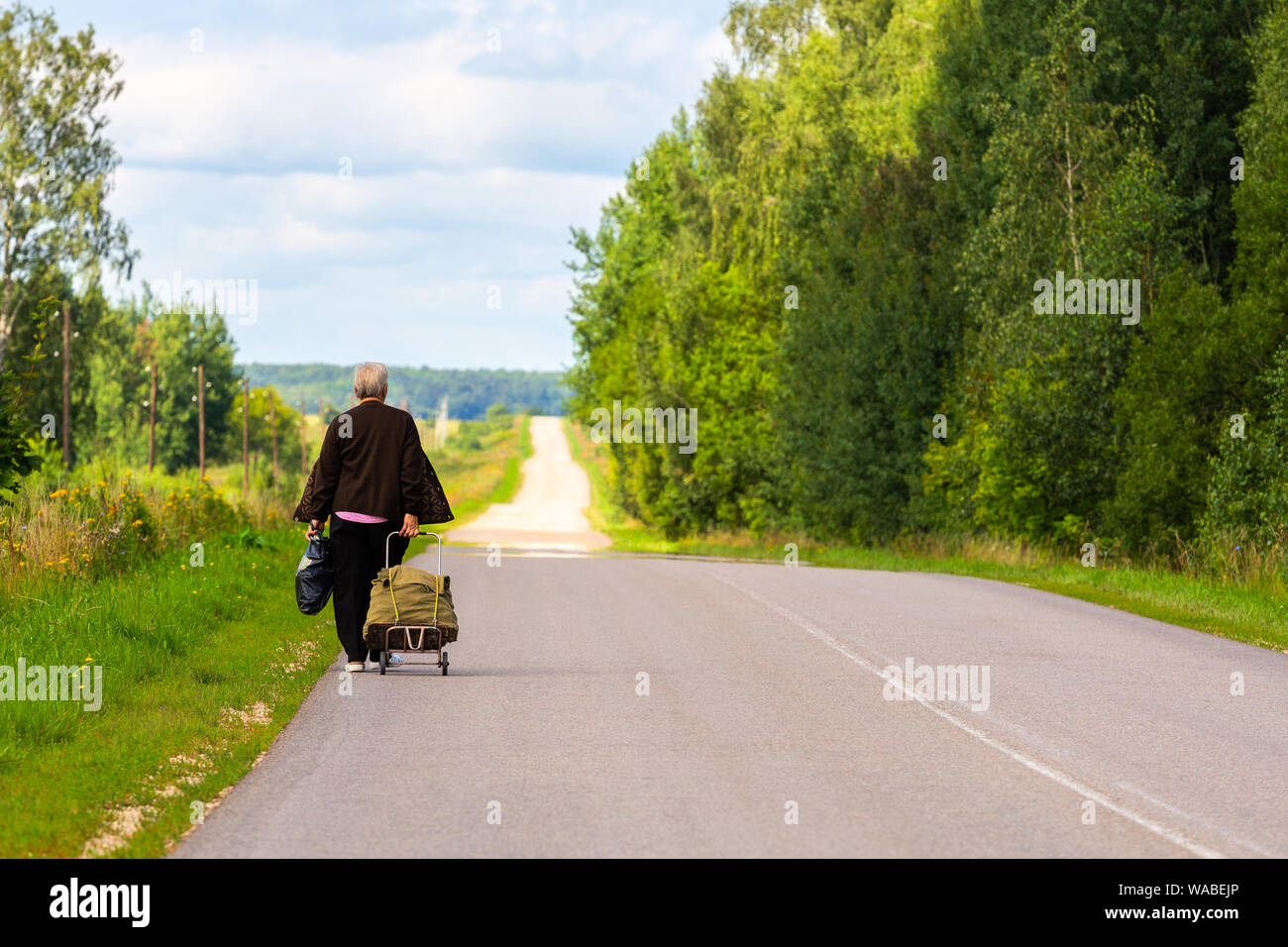 old woman with cart and black plastic bag walk away on side of rustic