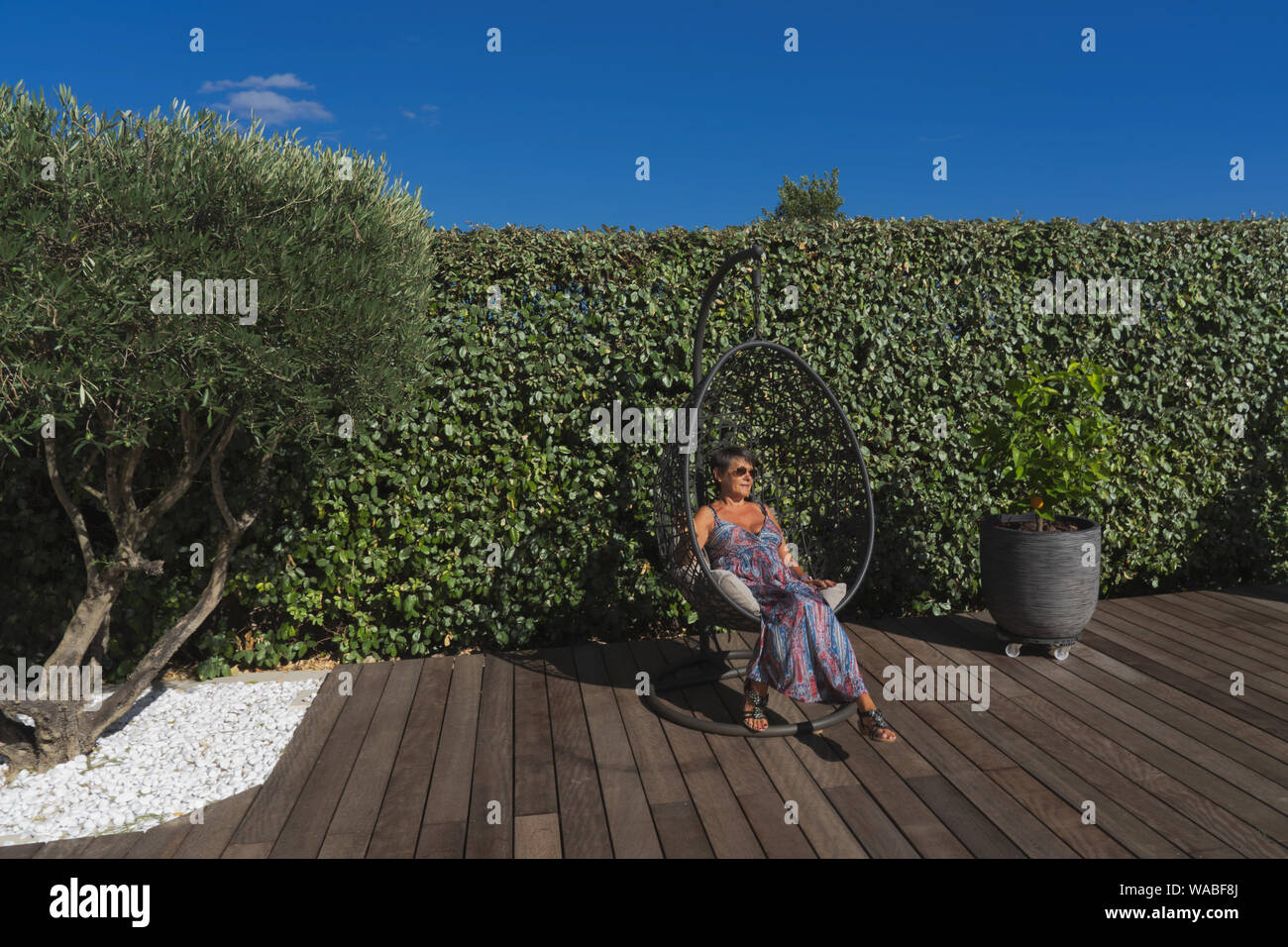 a woman sit in a suspended seat on a wooden floor in a garden with an olive tree Stock Photo