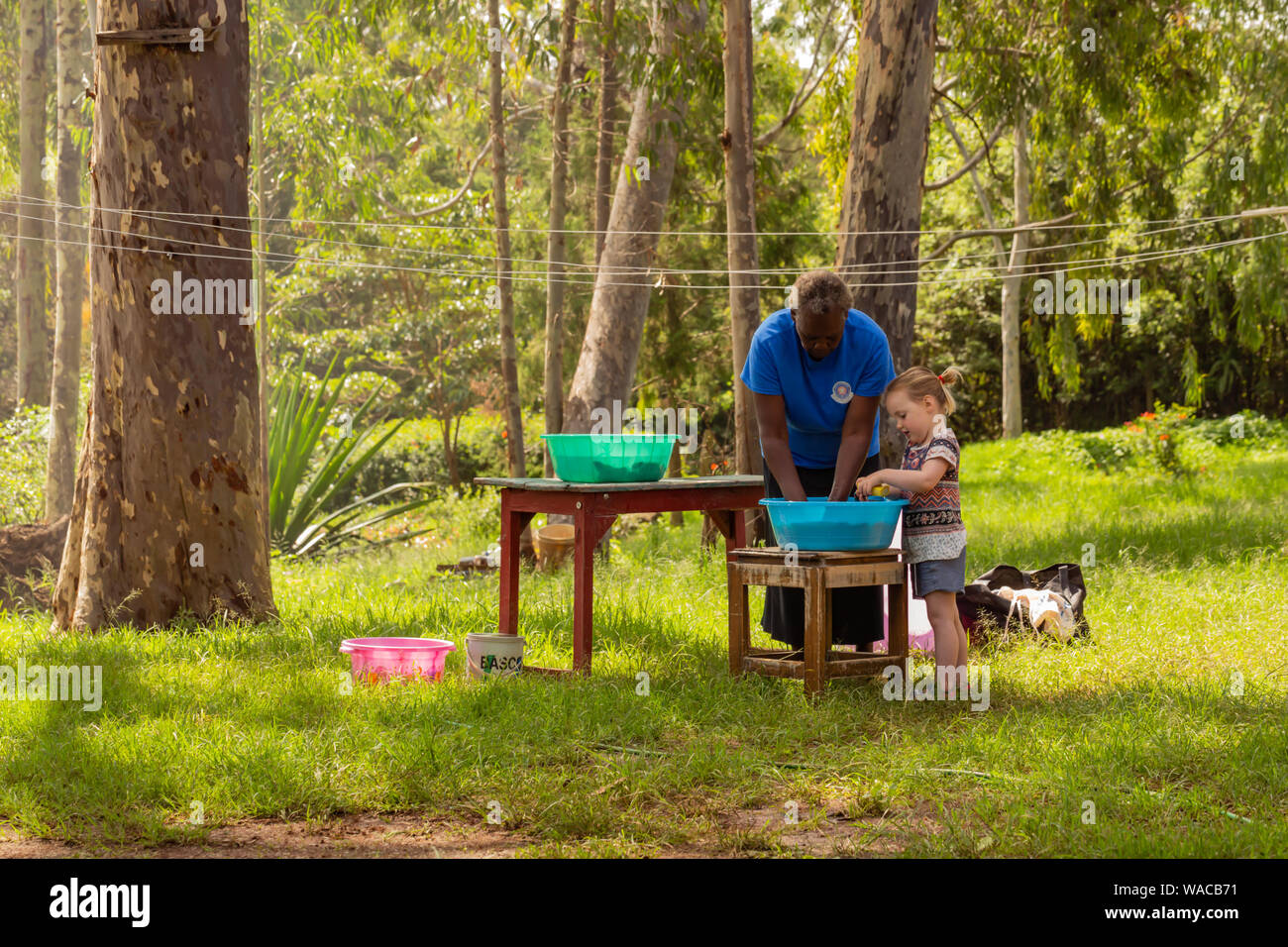 Nanyuki, Kenya – June 13th, 2019: Colour photograph of Kenyan woman teaching young white girl how to hand-wash laundry in beautiful garden. Stock Photo