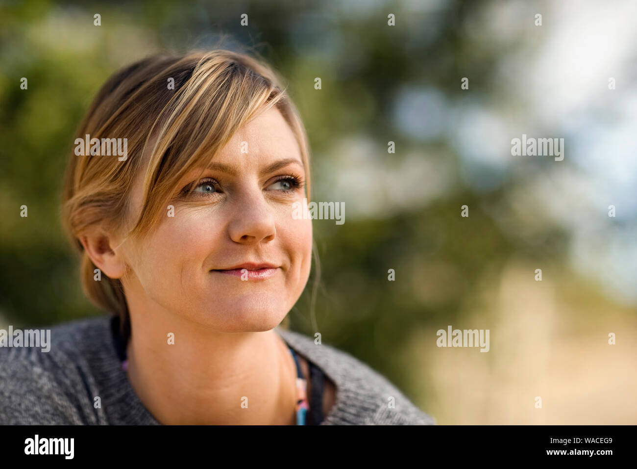 Smiling young woman looks thoughtfully into the distance as she sits outside. Stock Photo