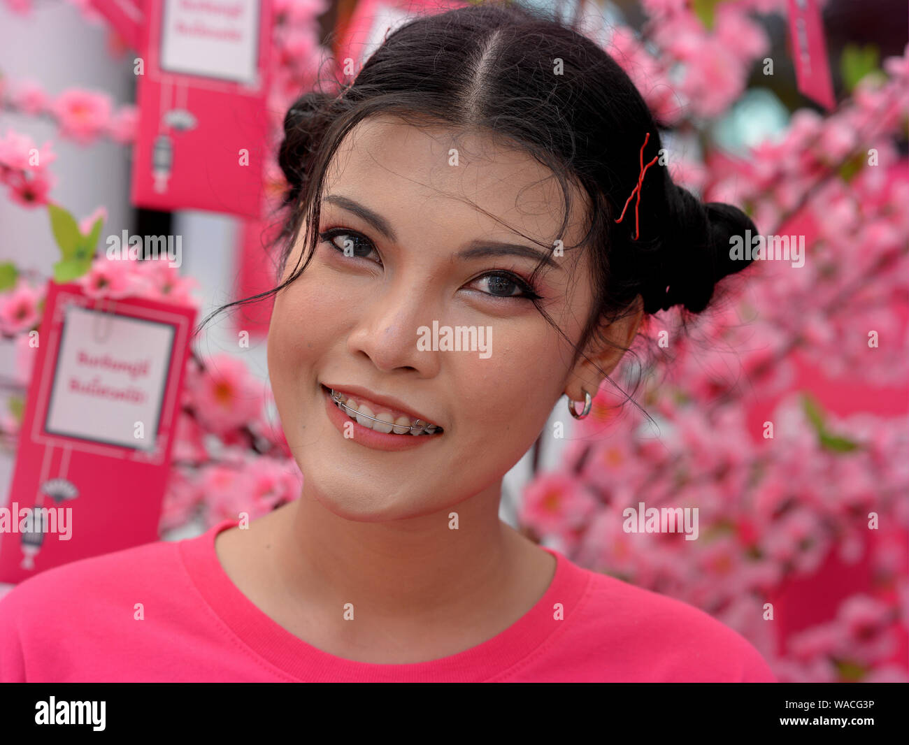 Young Thai beauty in pink poses for the camera in front of a pink blossom tree with attached Lunar New Year wishes at Chinese New Year 2019. Stock Photo