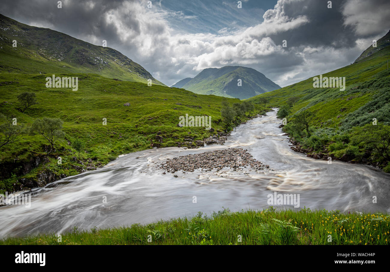 Green valley between beautiful mountain slopes, river with flowing water and dramatic sky at the Highlands of Scotland. Stock Photo