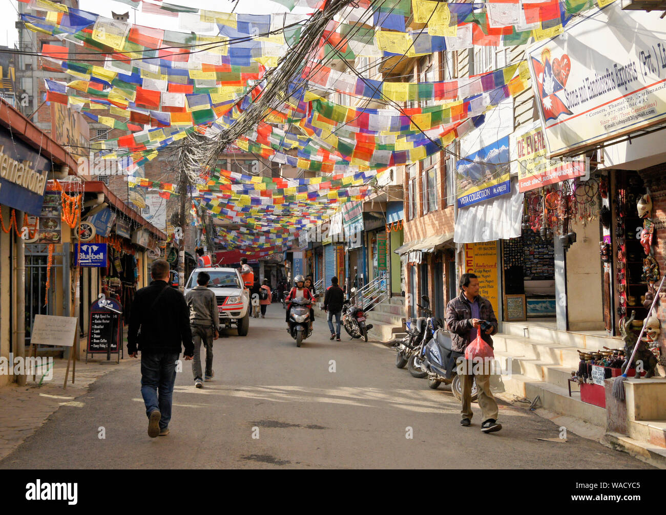 Street with Tibetan Buddhist prayer flags and electrical lines above, flanked by shops and