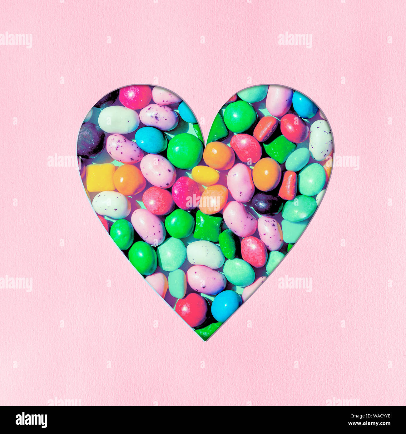 Heart shape filled with small colourful candies carved out of soft pink colored paper background. Sweetest day, birthday concept. Copy space for text. Stock Photo