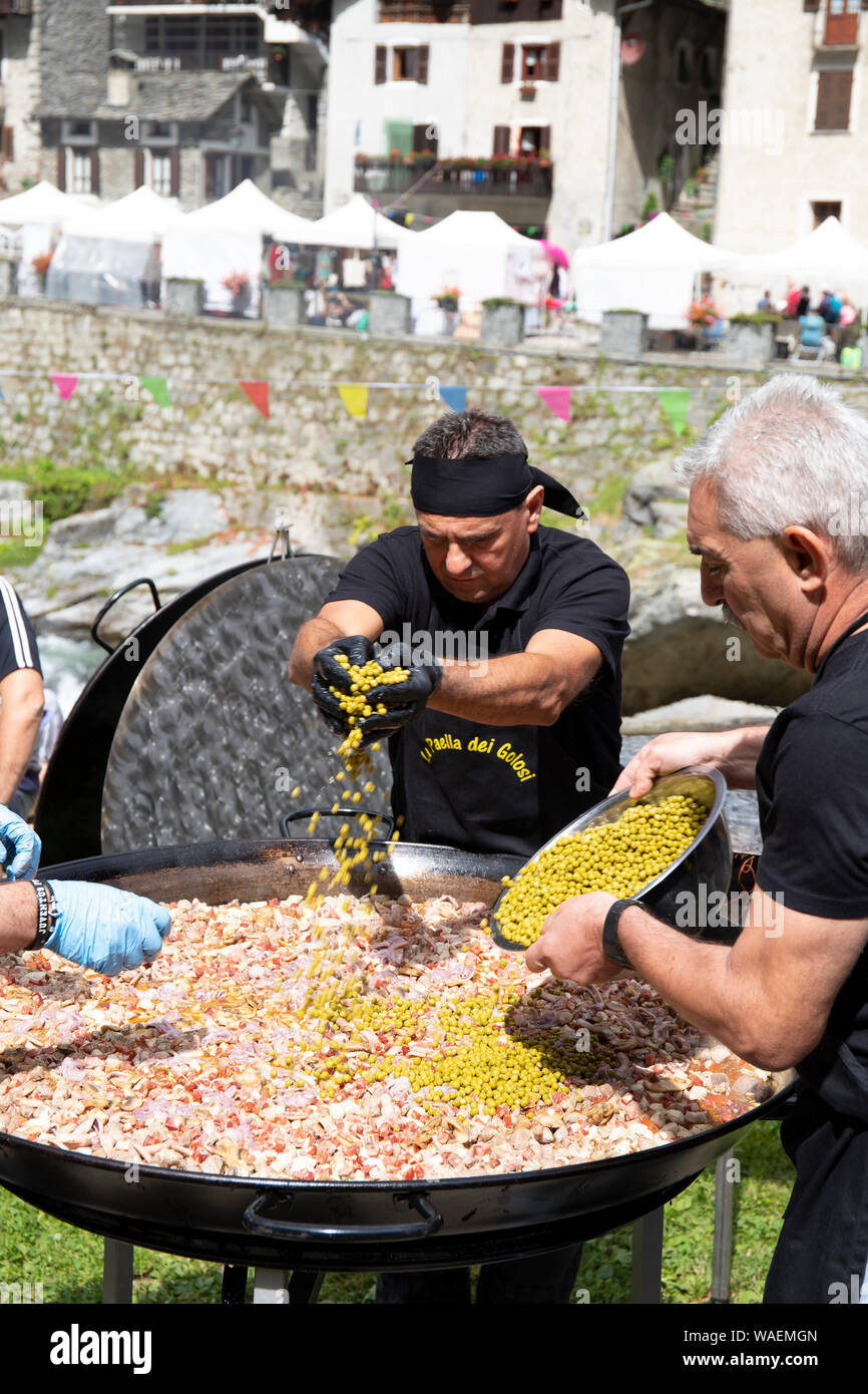 RASSA, ITALY - AUGUST 2019 - Men preparing paella in a big paellera, putting peas inside the pan. Rassa blueberry festival, Italy. Vertical shot. Stock Photo