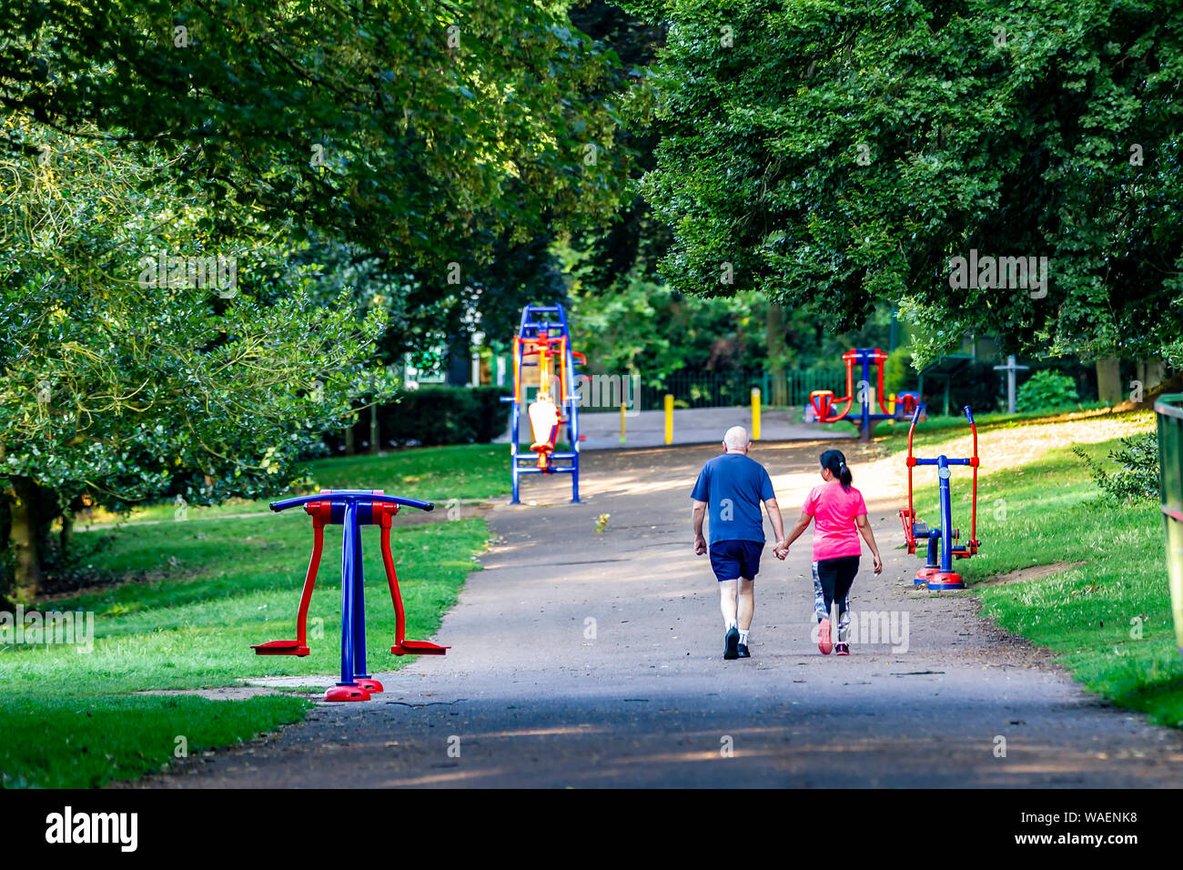 Northampton, UK. 2oth August 2019. Weather. A cool. sunny morning casting long shadows as this couple  walk up the towards Park Ave South  between the exercise machines during their morning exercise in Abington Park. Credit: Keith J Smith./Alamy Live News Stock Photo