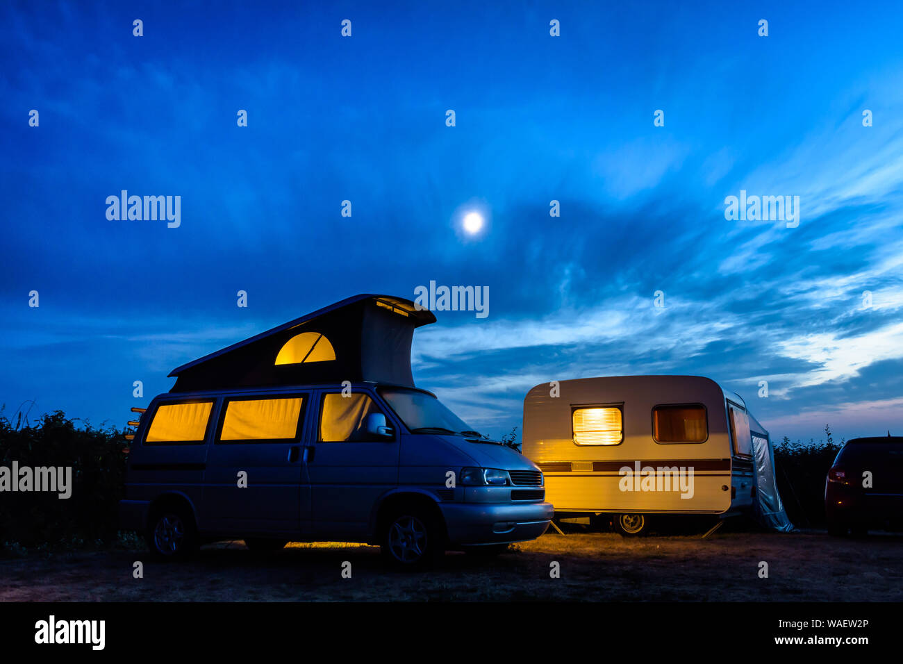 A small camper van parked next to a vintage caravan in a campsite, both illuminated from inside at nightfall with the moon glowing in a stormy sky. Stock Photo