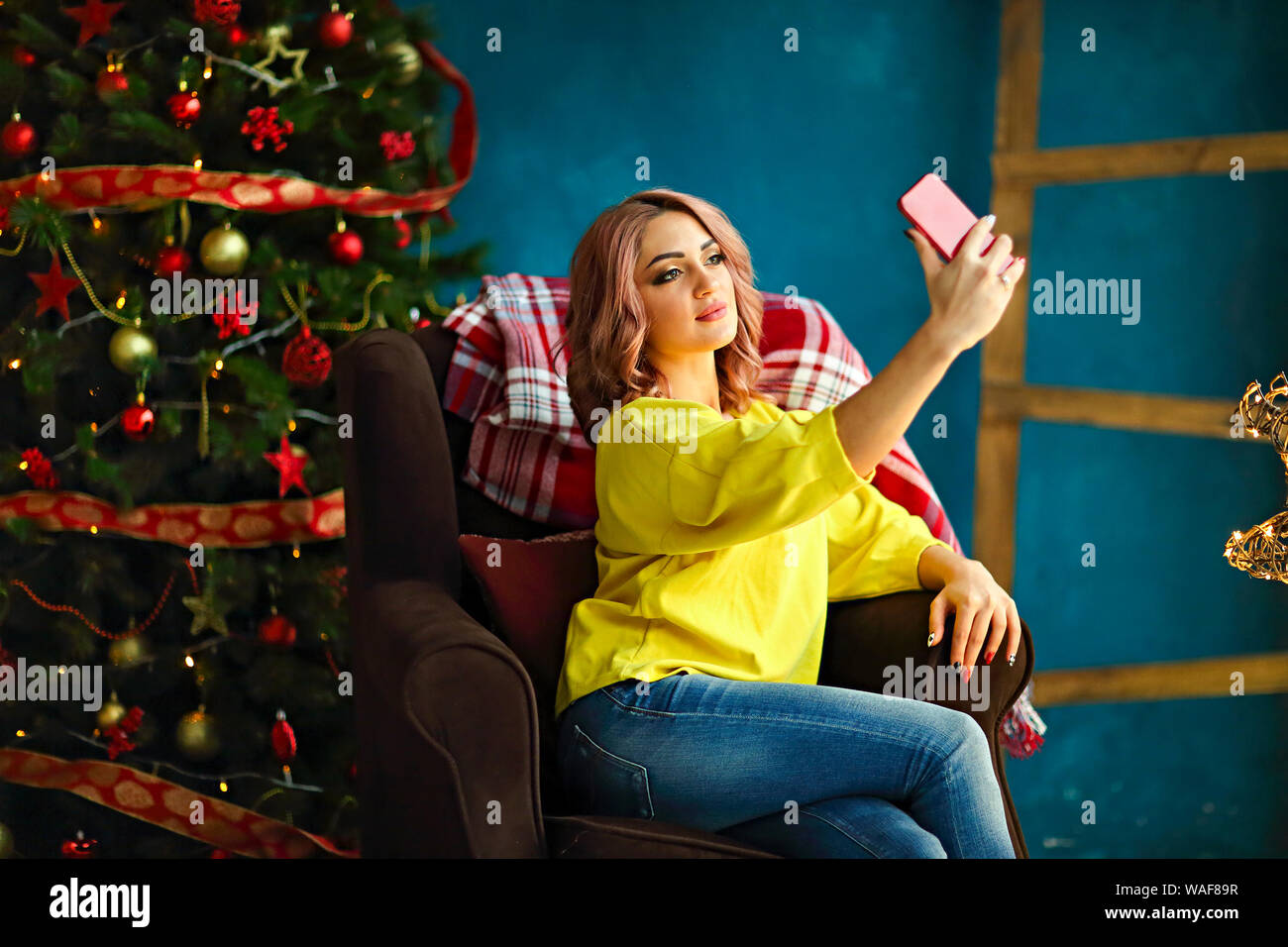 Young pretty woman taking selfie at home while sitting on armchair near Christmas tree Stock Photo
