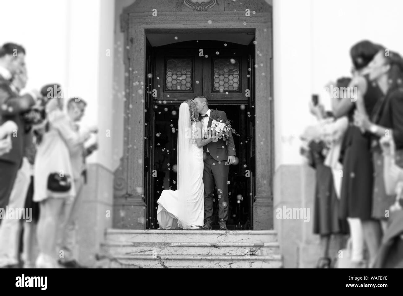 Newlyweds kissing while exiting the church after wedding ceremony, family and friends celebrating their love with the shower of soap bubbles, custom Stock Photo