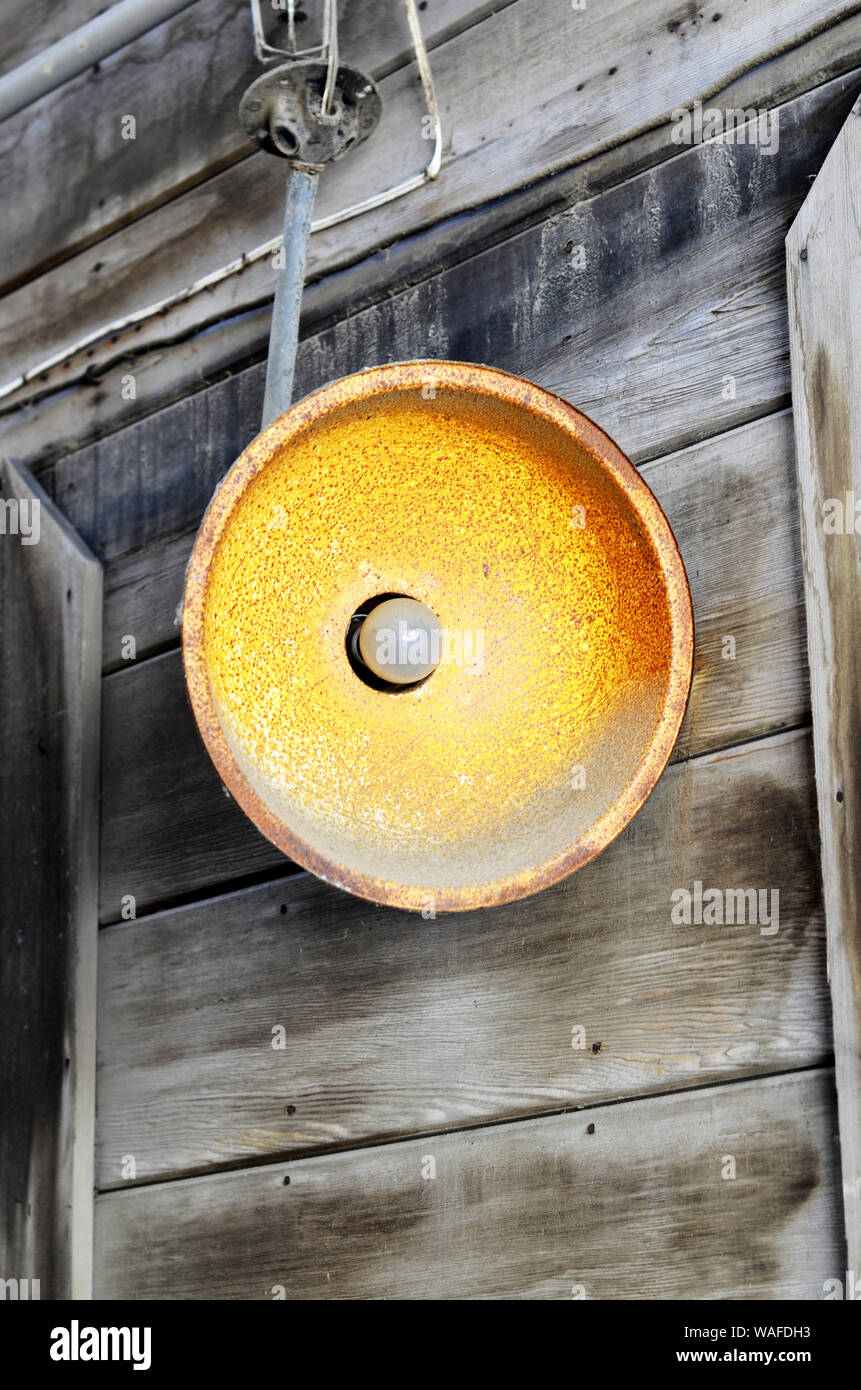 Rusted round open exterior light fixture with light bulb,  hanging in front of gray weathered wood siding in the background. Stock Photo