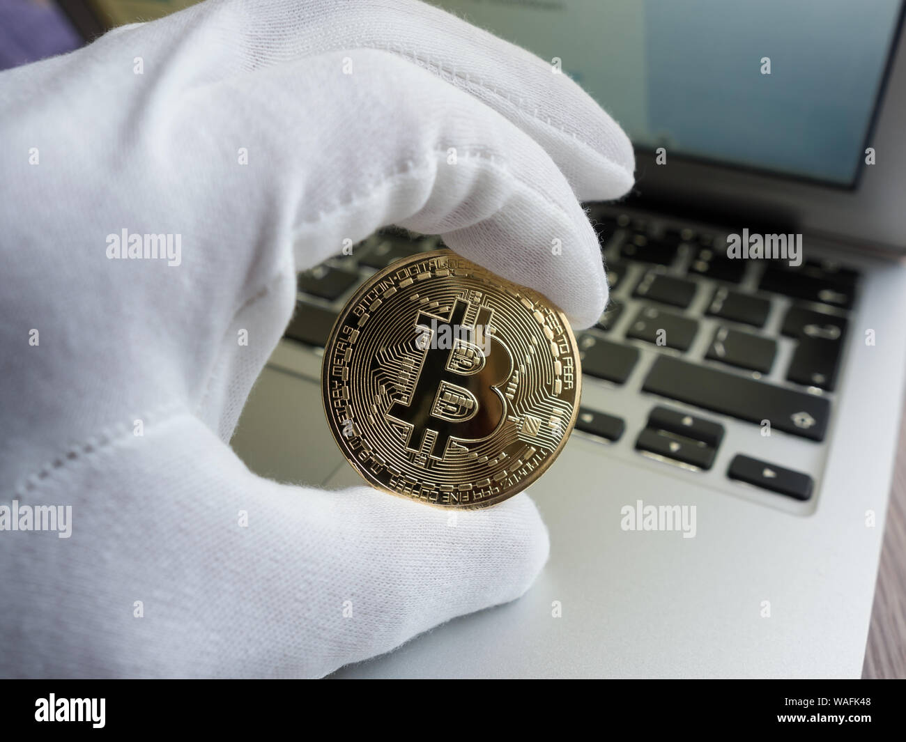 Men in white glove holding golden bitcoin against laptop. Focus on the coin. Stock Photo