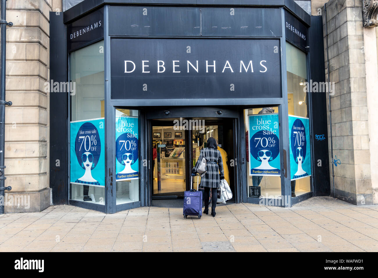 Back of Lady shopper carrying bags and wheeling a travel bag entering a Debenhams shop with large blue cross sale signs in the windows Princes Street Stock Photo